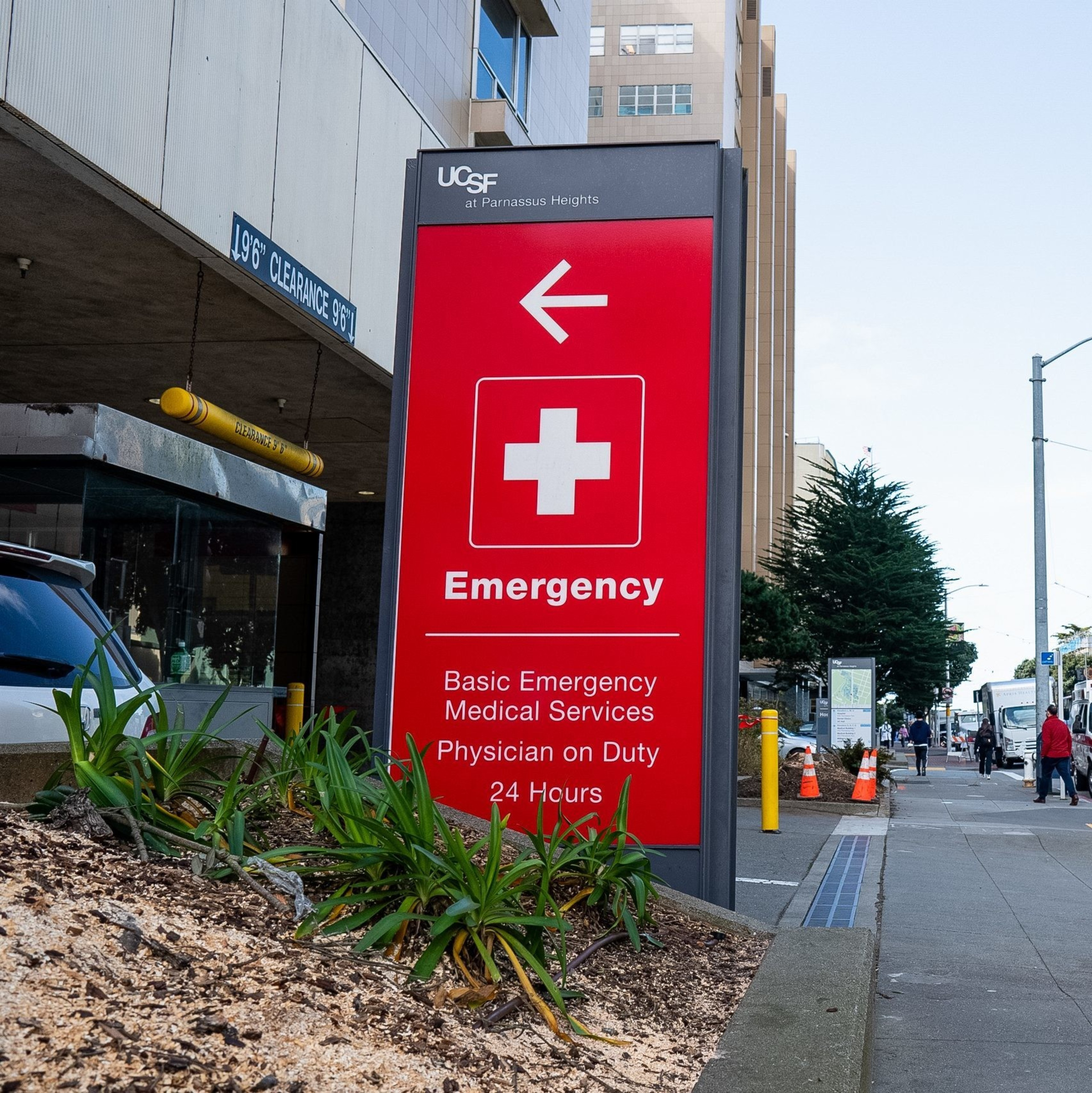 A red emergency sign with a white cross and arrow points left. The street has an ambulance, cars, pedestrians, and trees. Buildings line the background.