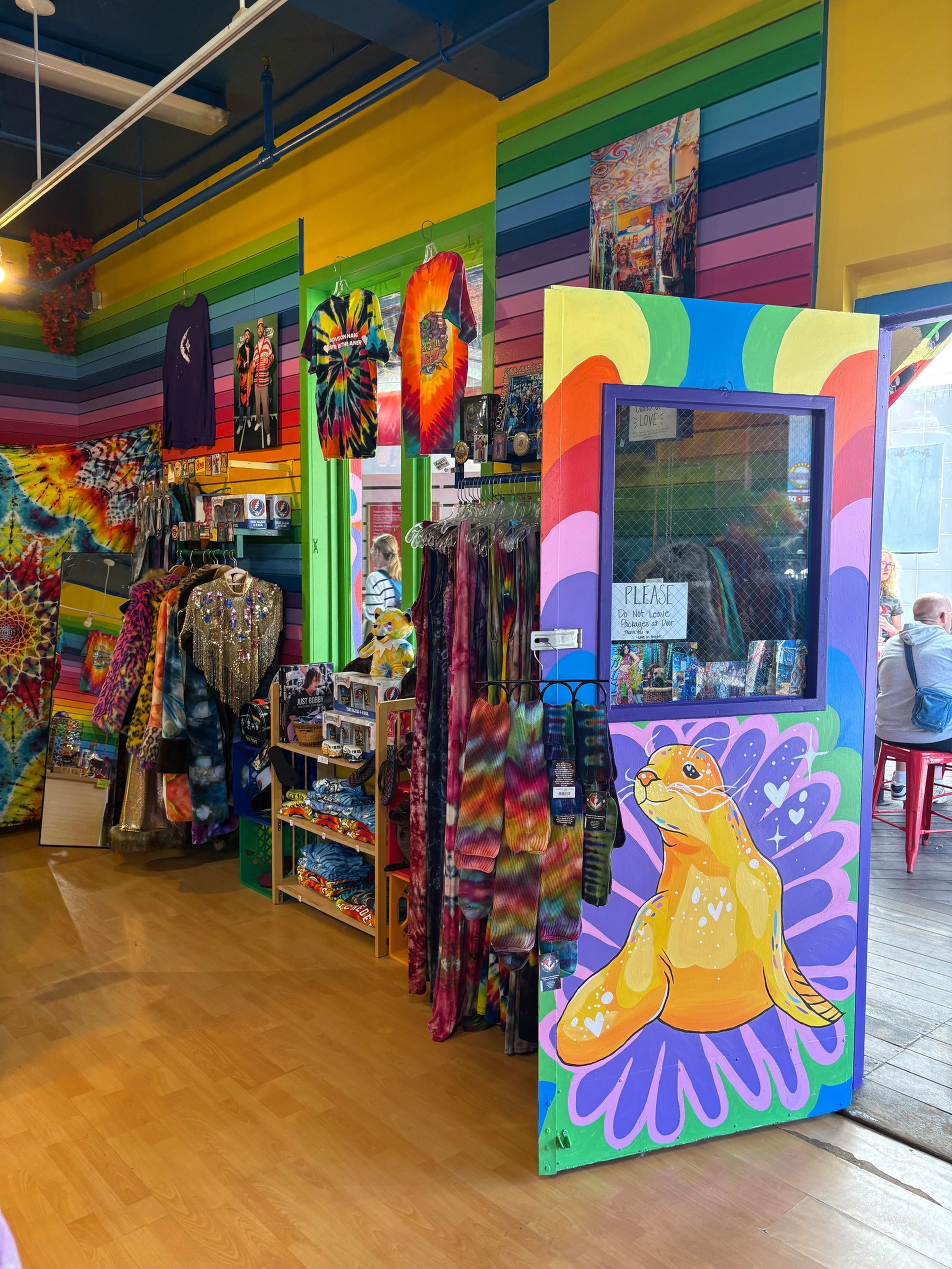 Colorful tie-dye clothes and accessories hang and fold inside a rainbow-striped shop with a door painted with a bright seal and purple flower design.