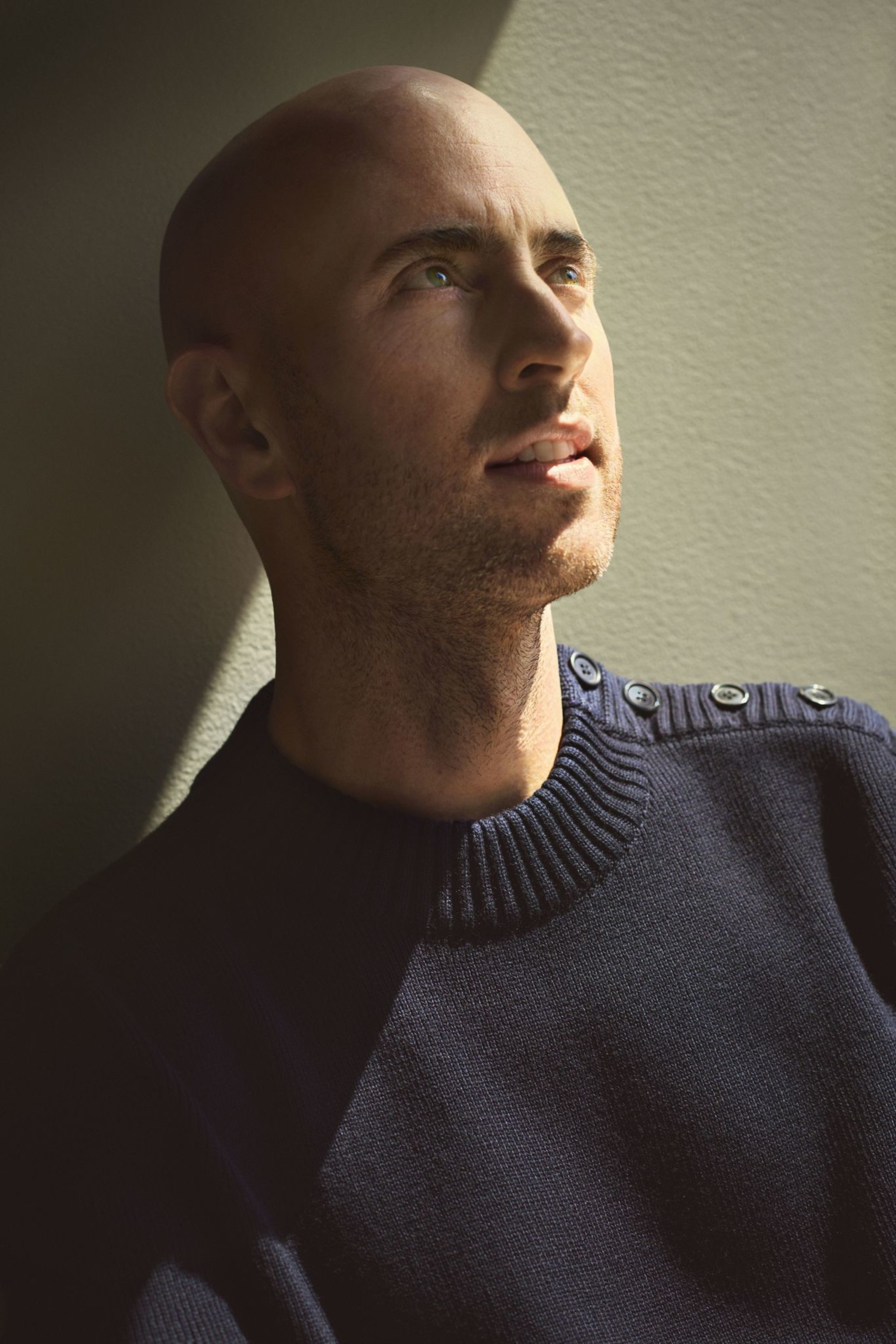 A bald man with light skin gazes upward toward a soft light, wearing a dark navy sweater with buttons on the shoulder against a plain beige wall.