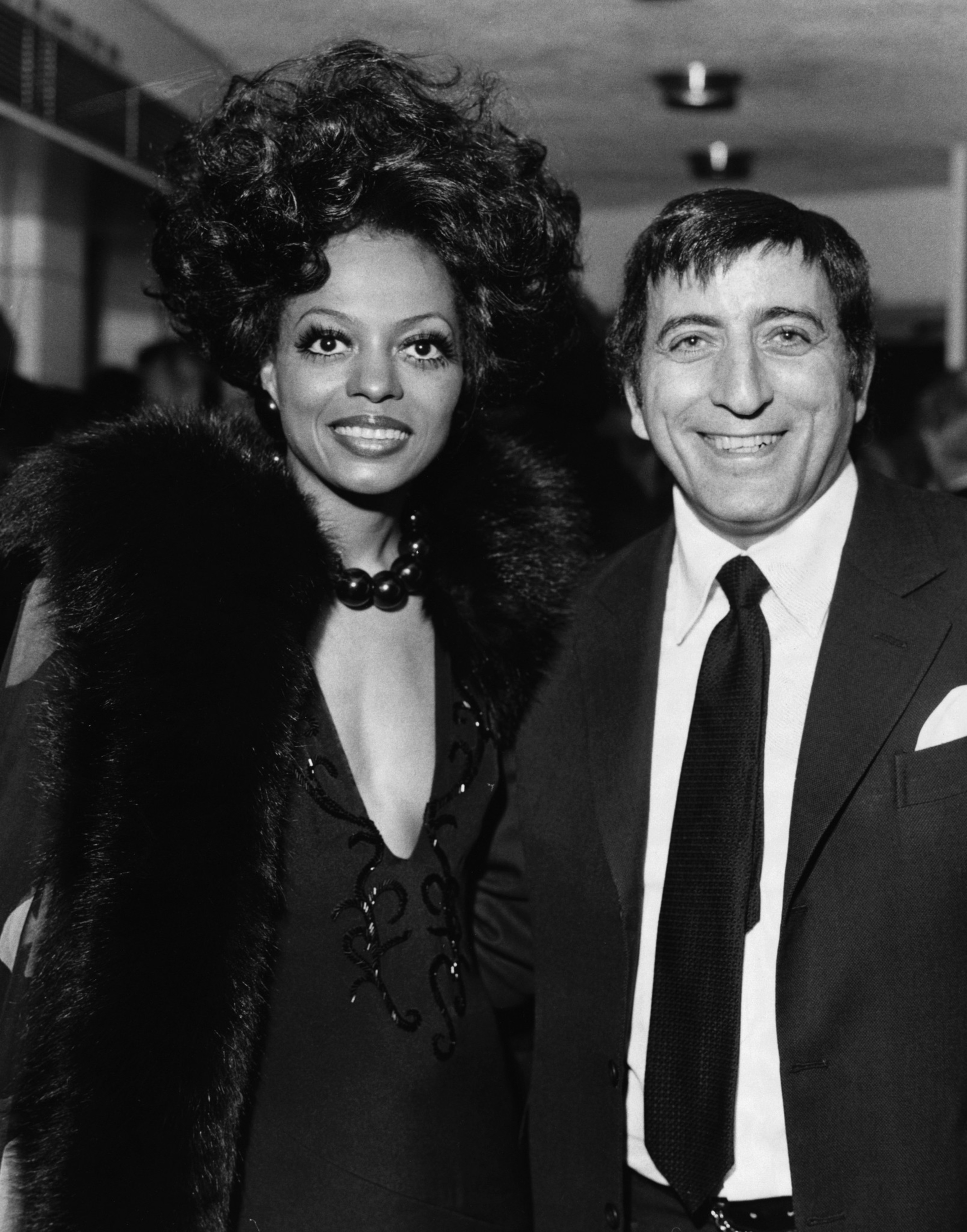A woman with voluminous hair and a fur stole stands next to a smiling man in a dark suit and tie, both dressed formally.