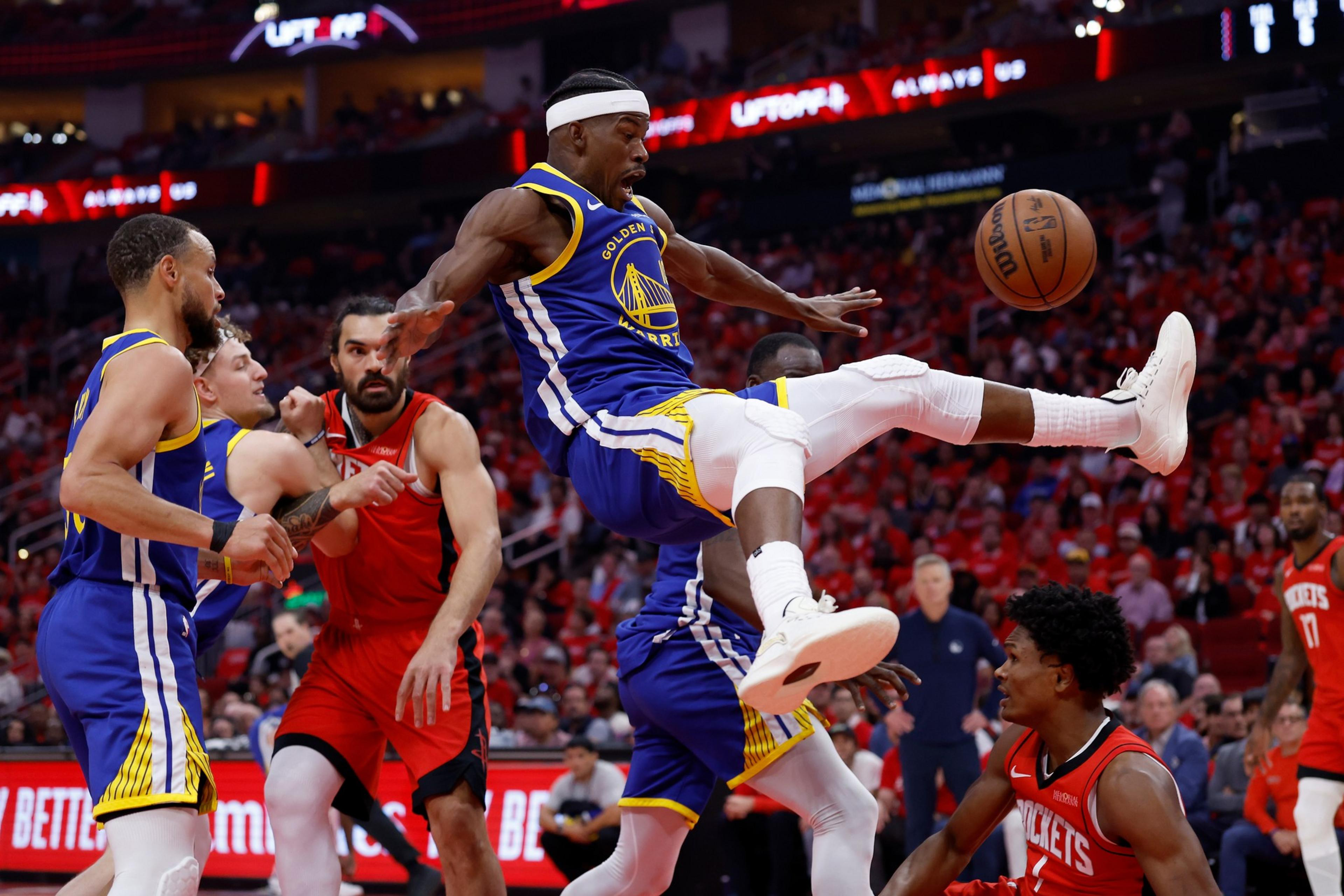 A basketball player in a blue and yellow uniform leaps with legs spread, attempting to control the ball. Opponents in red jerseys watch closely.