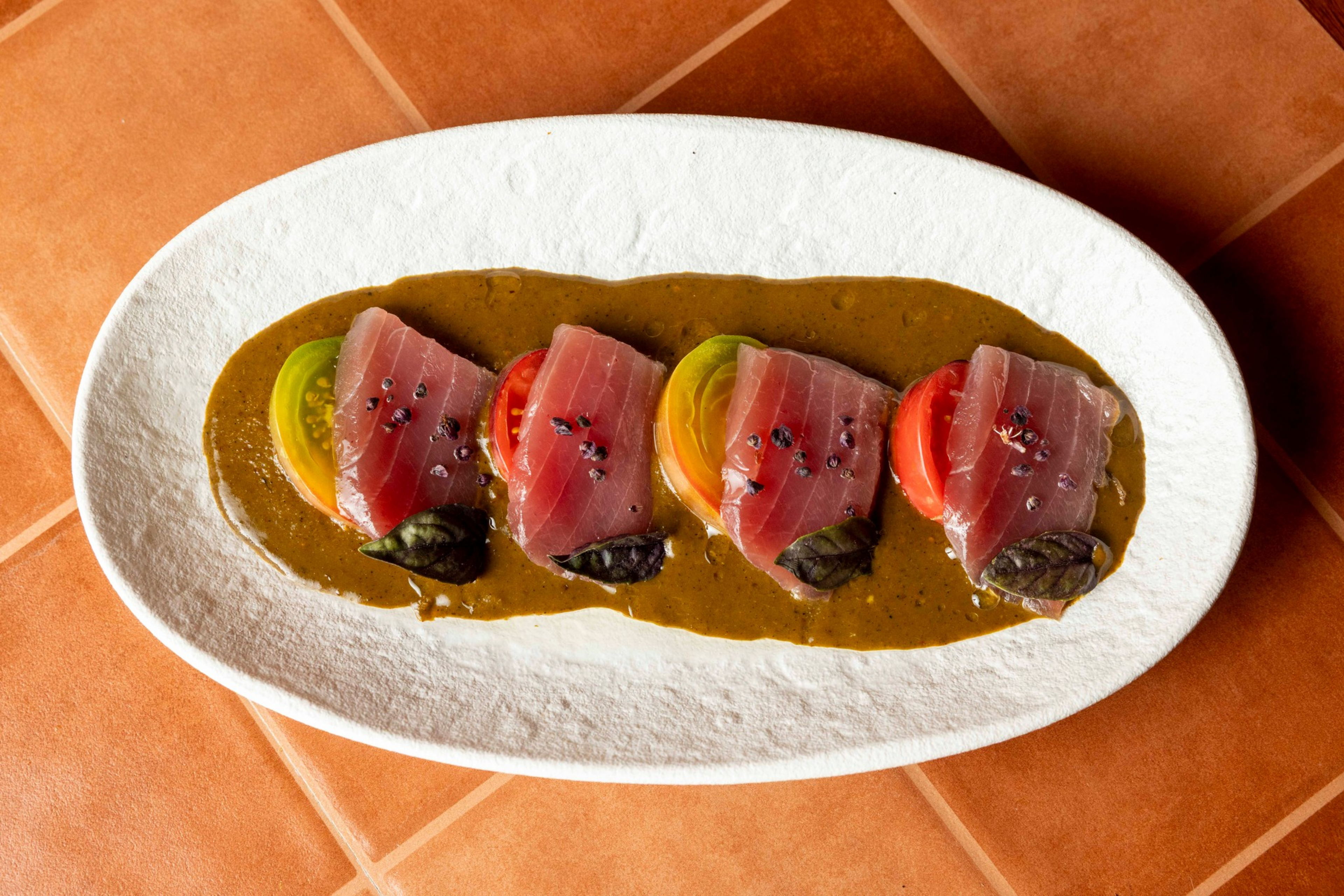 Four pieces of raw tuna sit on sliced red and yellow tomatoes, garnished with dark green leaves and pepper, all on a brown sauce on a white plate.