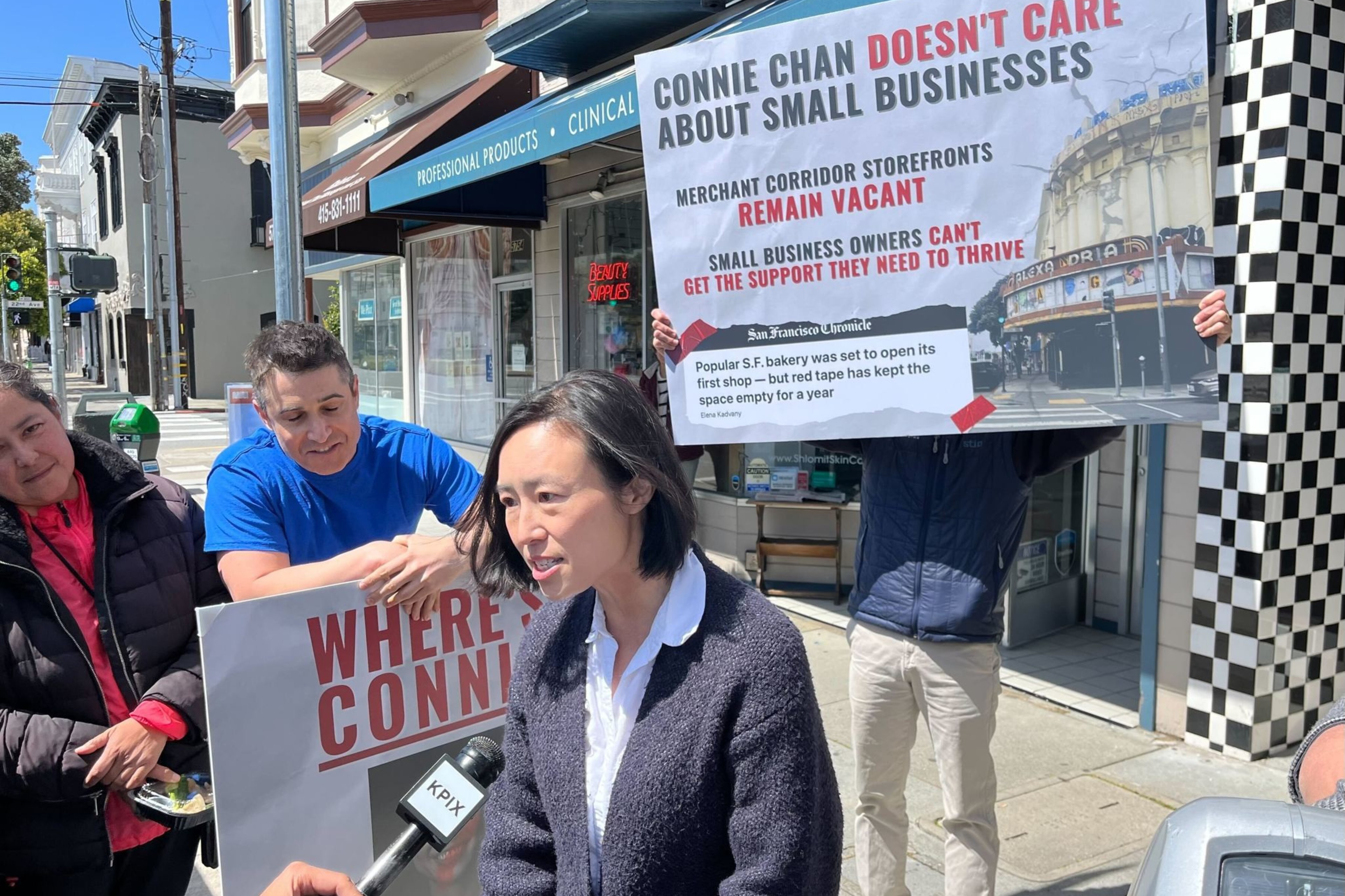 A person is being interviewed on a sunny street, holding a sign about local business concerns, with others holding a larger banner.
