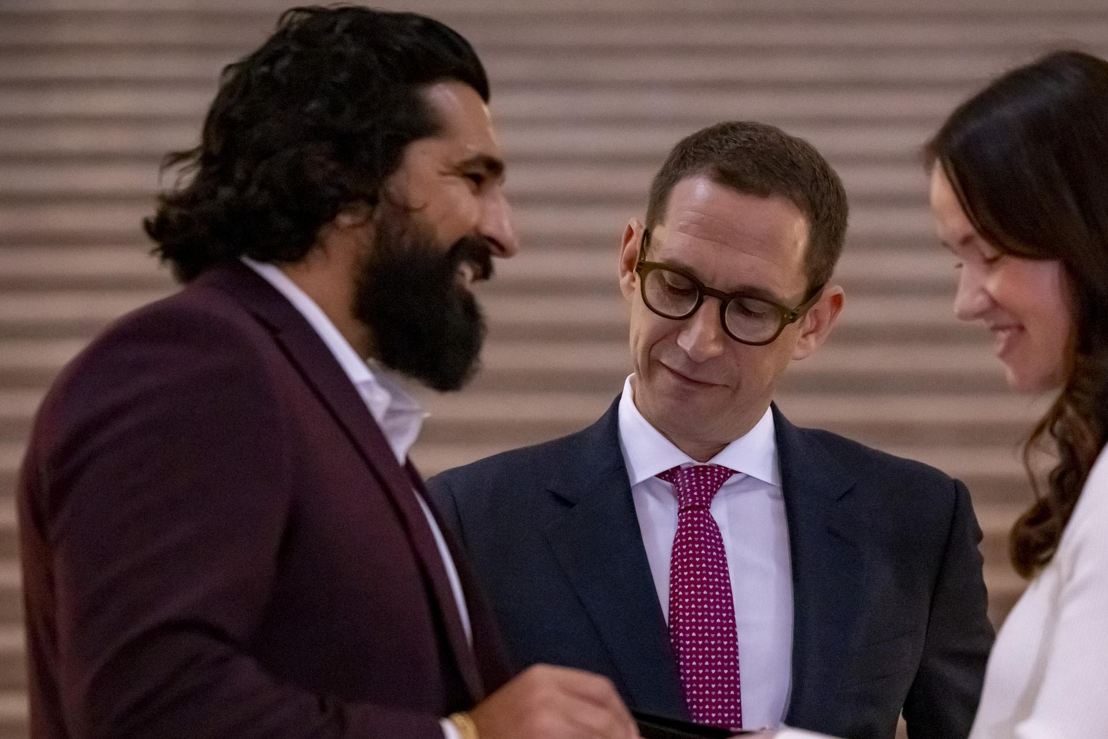 A couple is exchanging rings in a ceremony while a man, possibly an officiant, watches them approvingly. They are smiling and dressed formally.