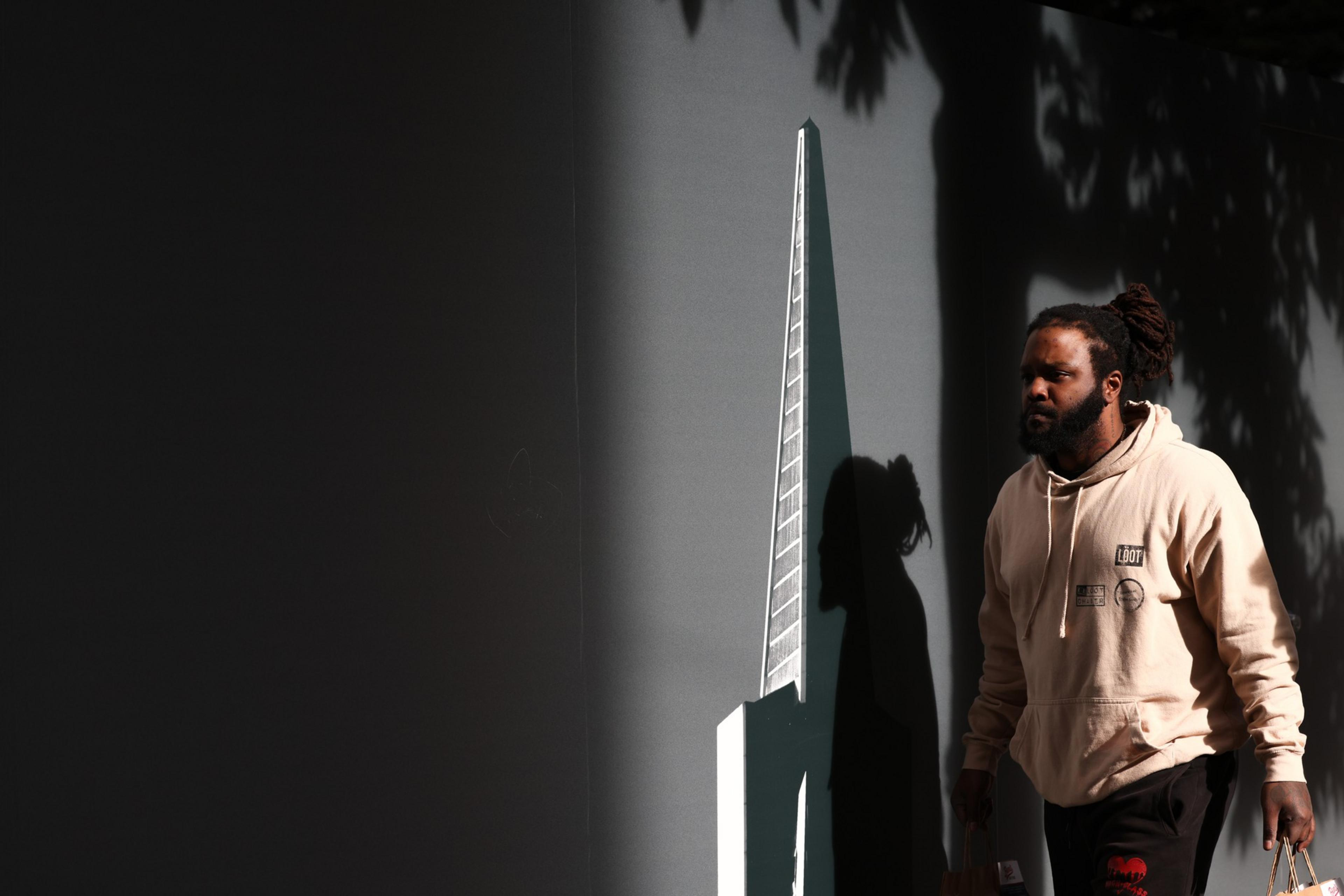 A man with dreadlocks wearing a light beige hoodie walks past a wall with a triangular architectural model and his shadow cast on it.