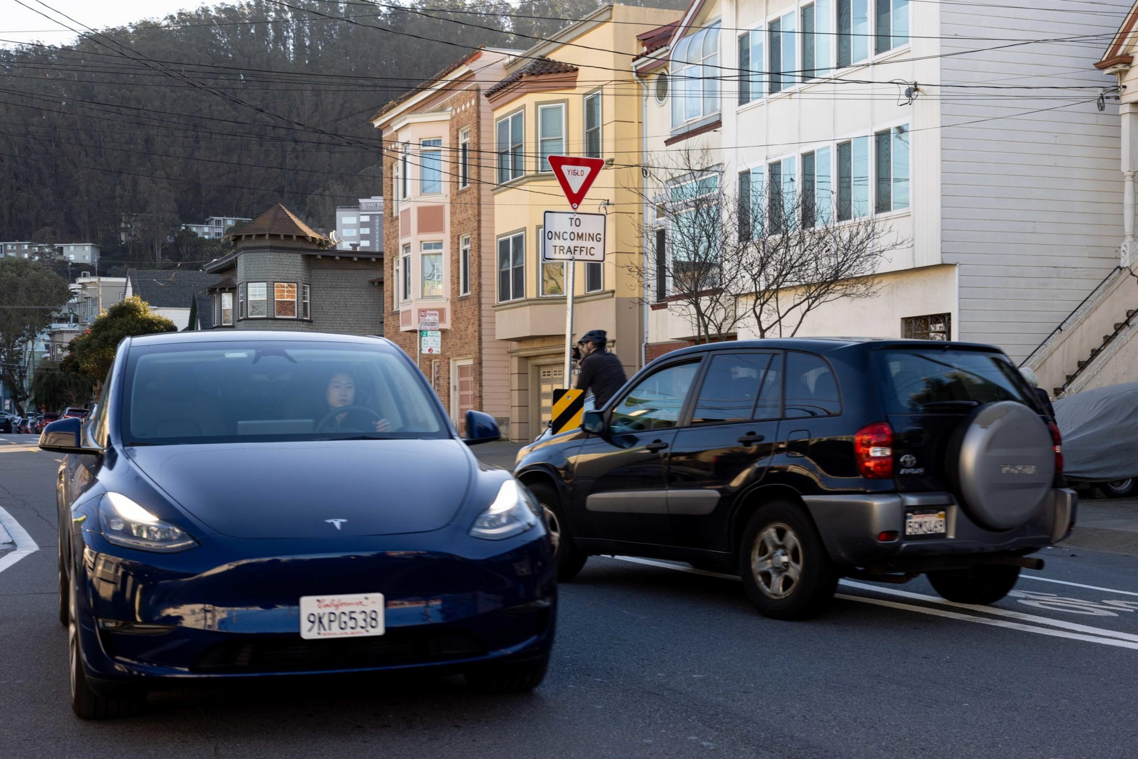 A blue Tesla and a black SUV are at an intersection with a &quot;Yield to Oncoming Traffic&quot; sign. A cyclist is nearby, and residential buildings are in the background.
