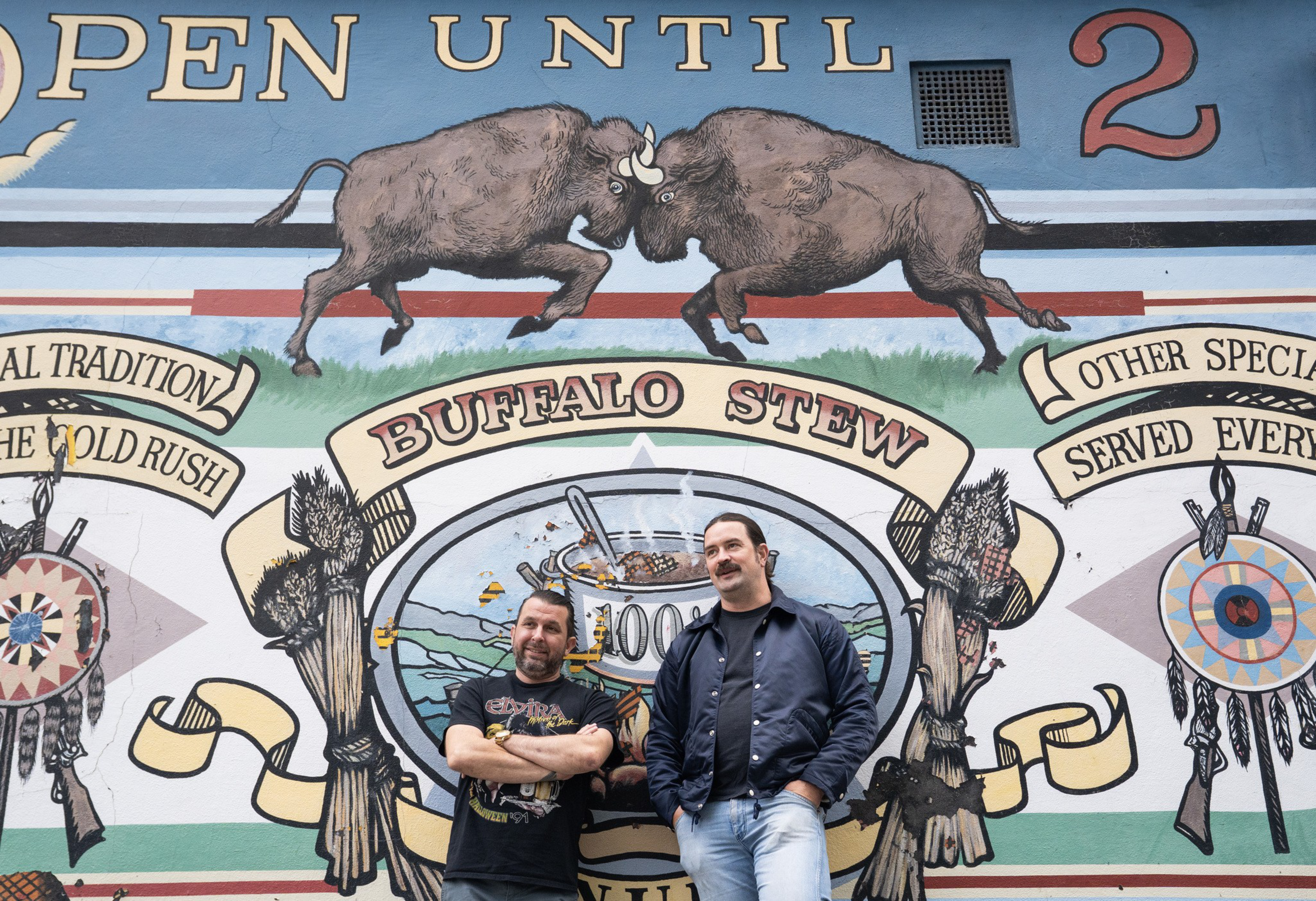 Two men stand in front of a colorful mural featuring two buffalo head-butting above a banner that reads "BUFFALO STEW" with other decorative elements.