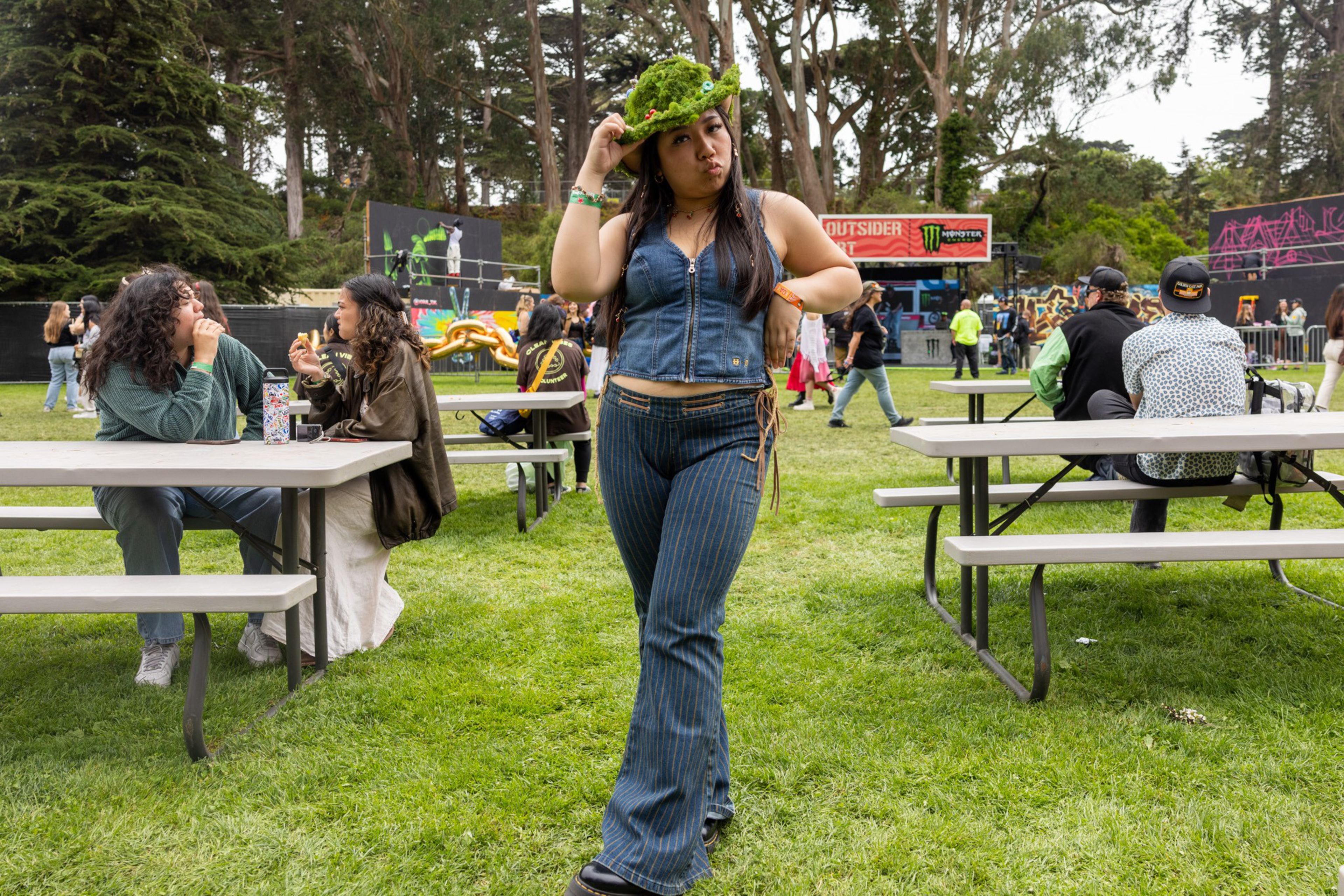 A woman wearing a moss-covered hat, denim vest, and striped pants stands on grass, surrounded by people sitting at picnic tables in an outdoor park setting.