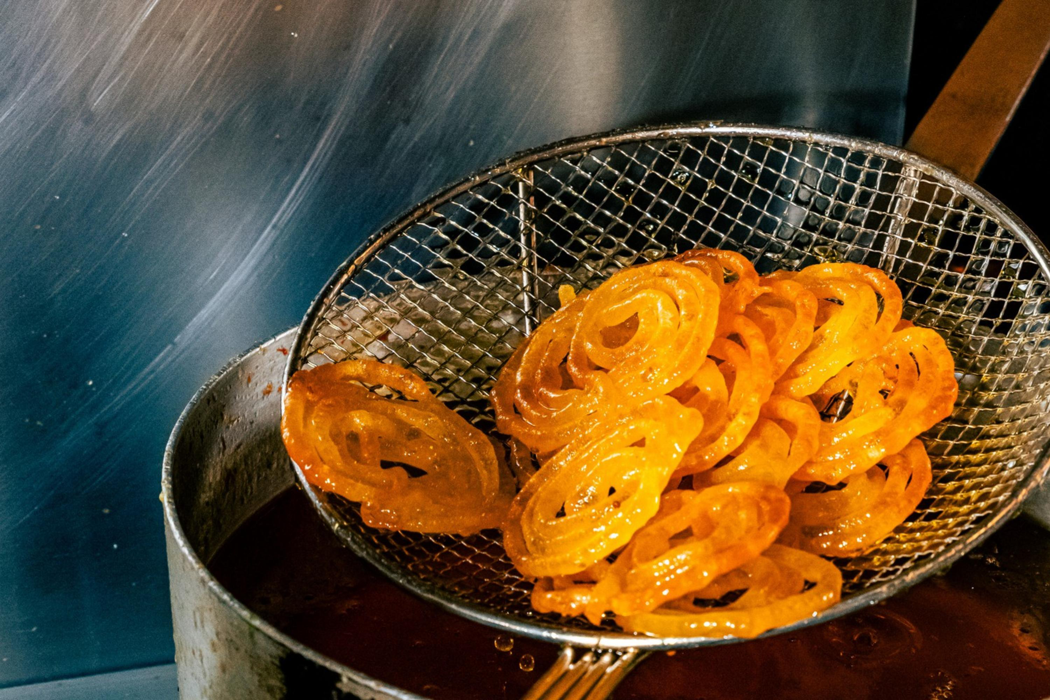 Golden, spiral-shaped jalebi sweets are being lifted from hot oil with a wire skimmer over a metal pot.