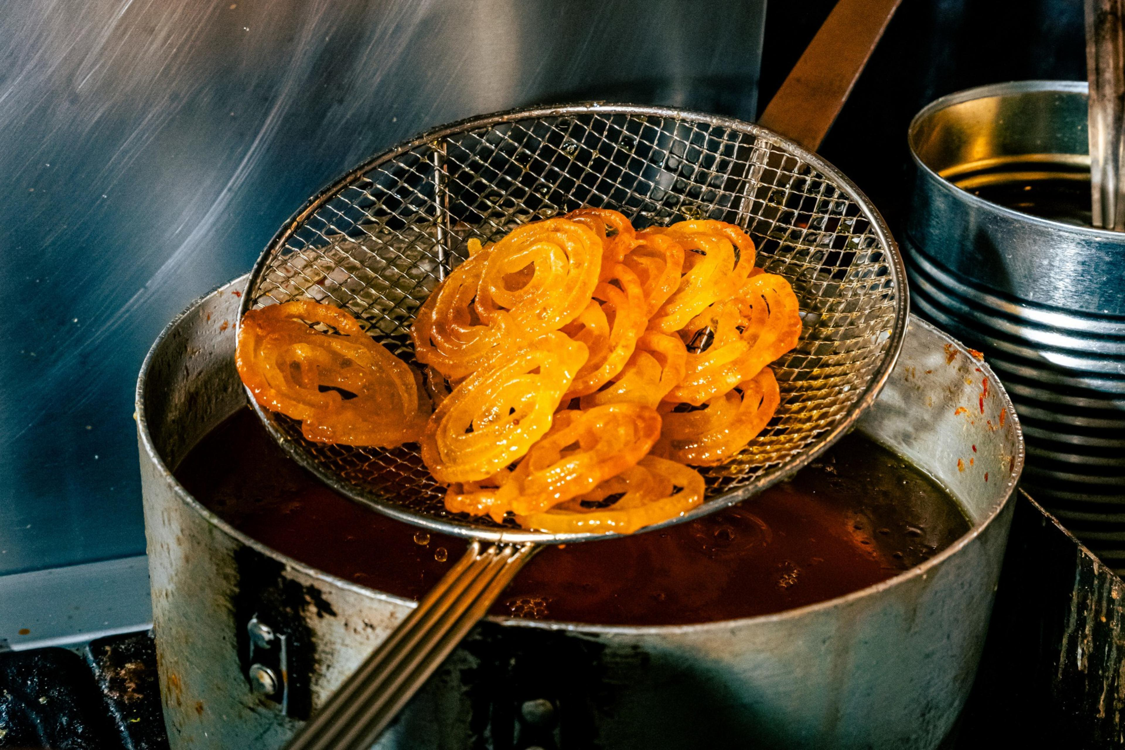 Golden, spiral-shaped jalebi sweets are being lifted from hot oil with a wire skimmer over a metal pot.