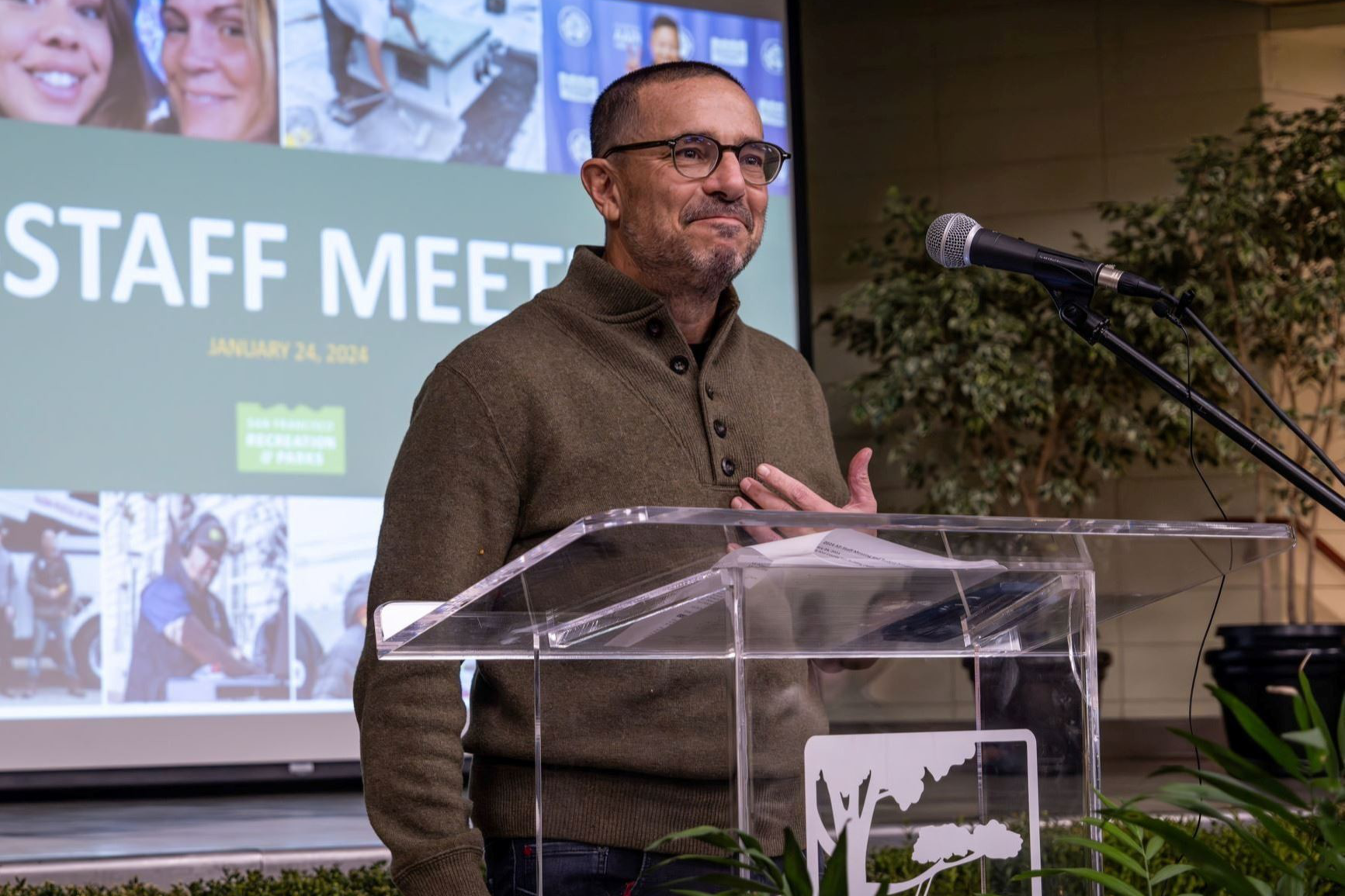 A man in glasses and a brown sweater stands behind a clear podium with a microphone, speaking at a staff meeting with a green and white presentation screen behind him.