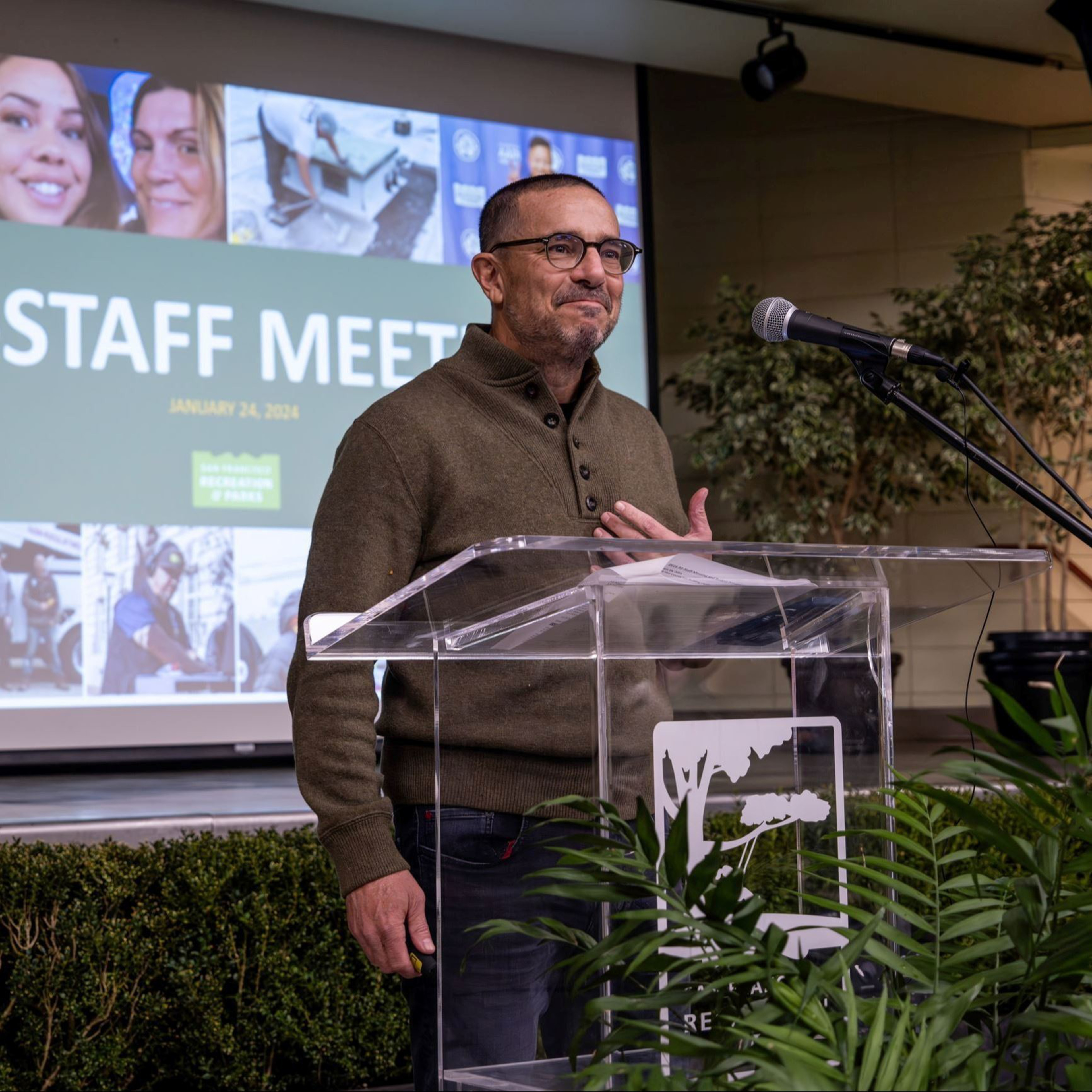 A man in glasses and a brown sweater stands behind a clear podium with a microphone, speaking at a staff meeting with a green and white presentation screen behind him.