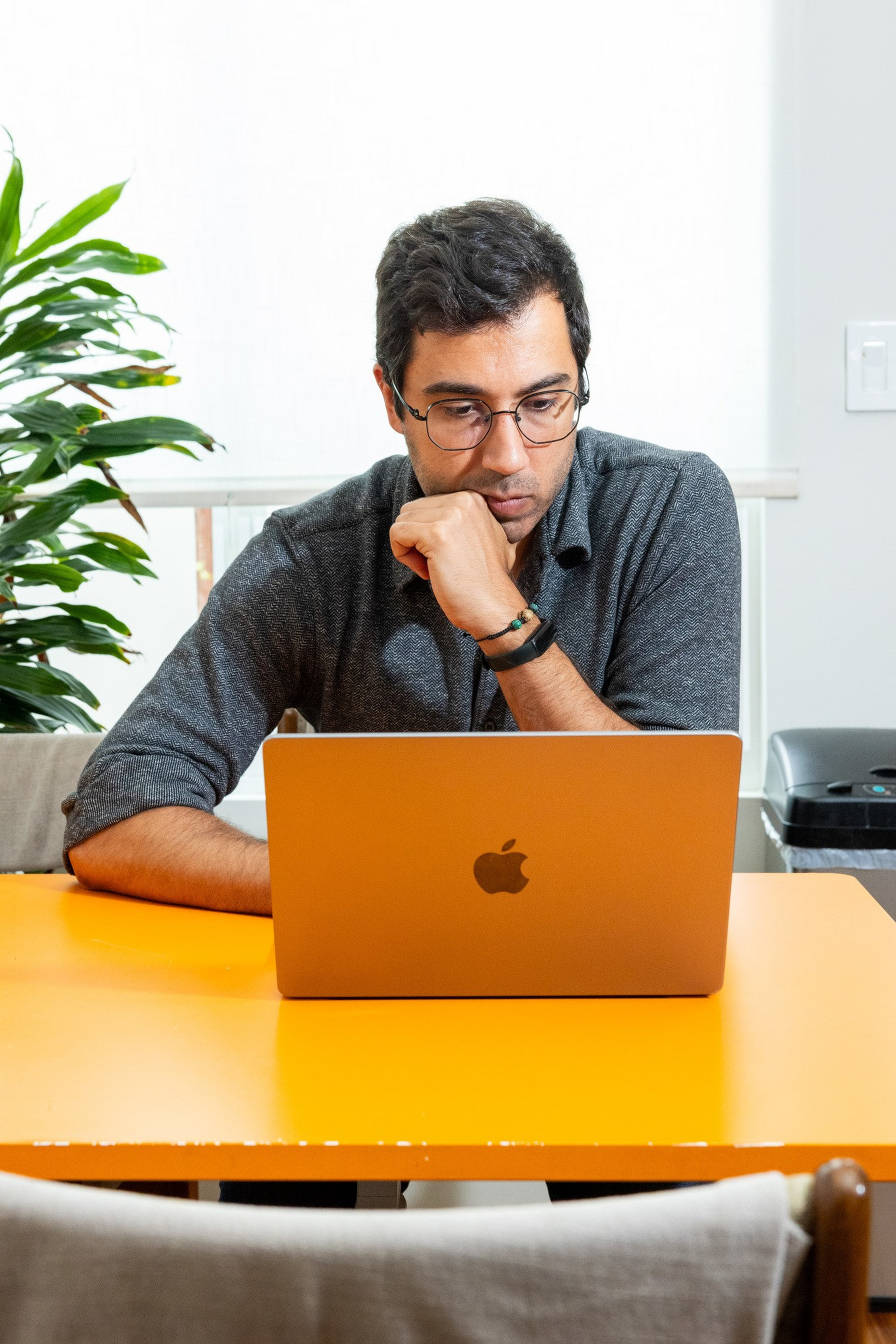 A man with glasses is focused on a laptop at a bright orange table, resting his chin on his hand, with a plant and a trash bin in the background.