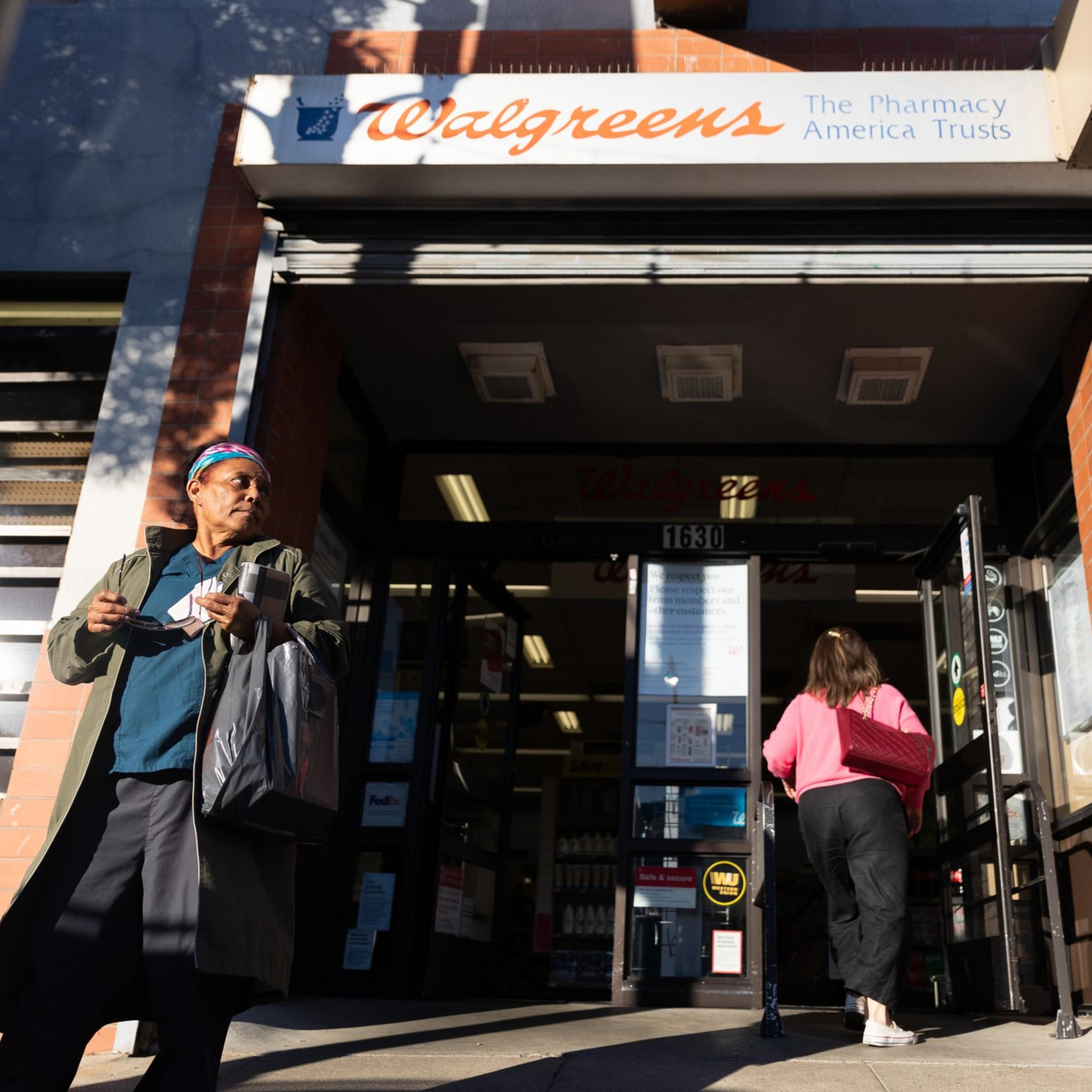 The image shows a Walgreens entrance with a person walking out holding a bag and another person entering the store. The building is brick with visible signage.