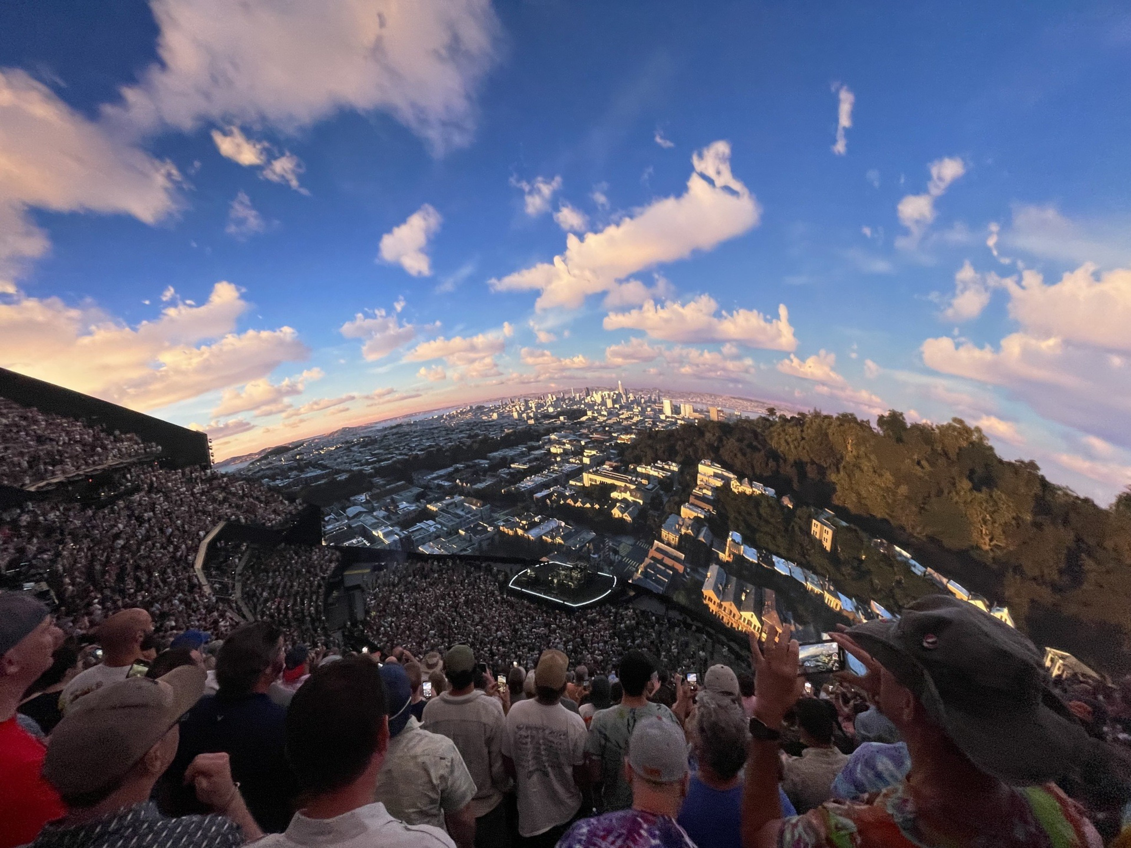 A large crowd is attending an outdoor concert at dusk with the city skyline and colorful sky in the distance, creating a vibrant and dynamic urban scene.