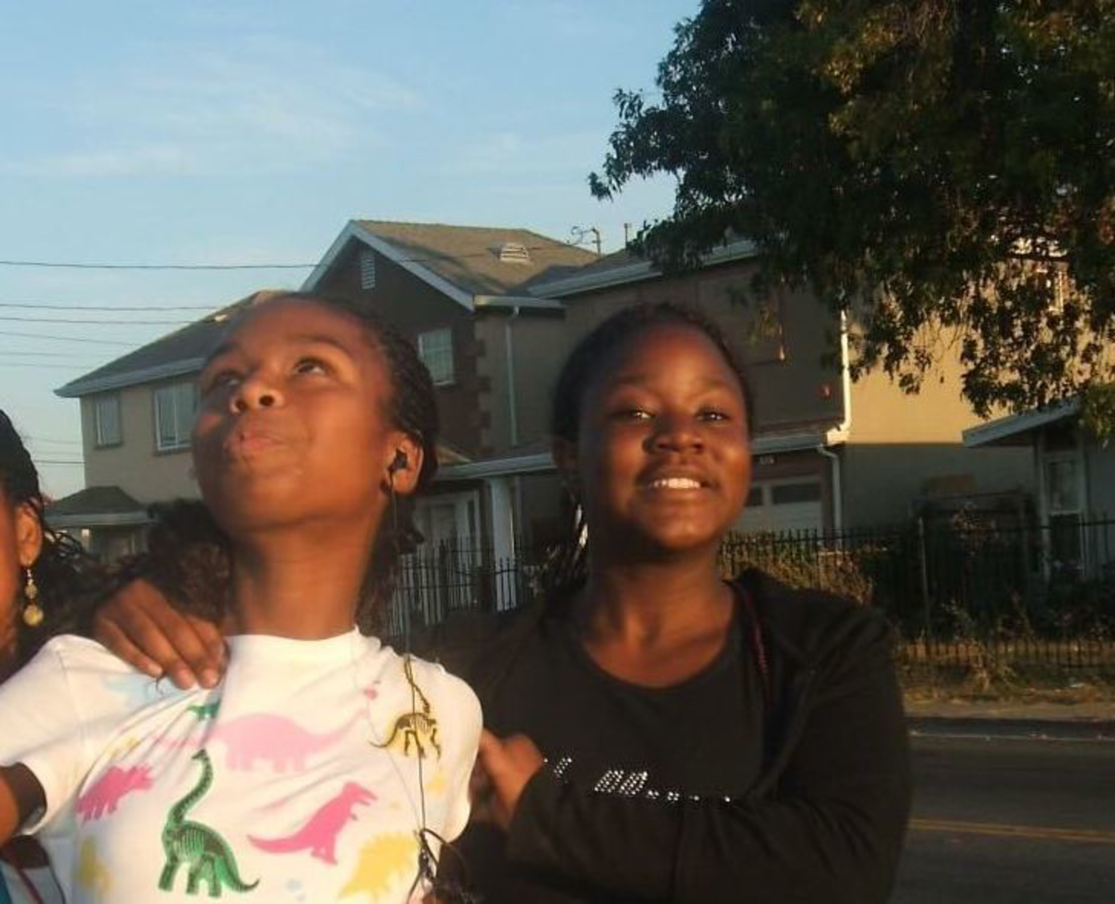Two girls stand outdoors in front of houses, one wearing a white dinosaur-themed shirt looking up, and the other in black smiling at the camera.