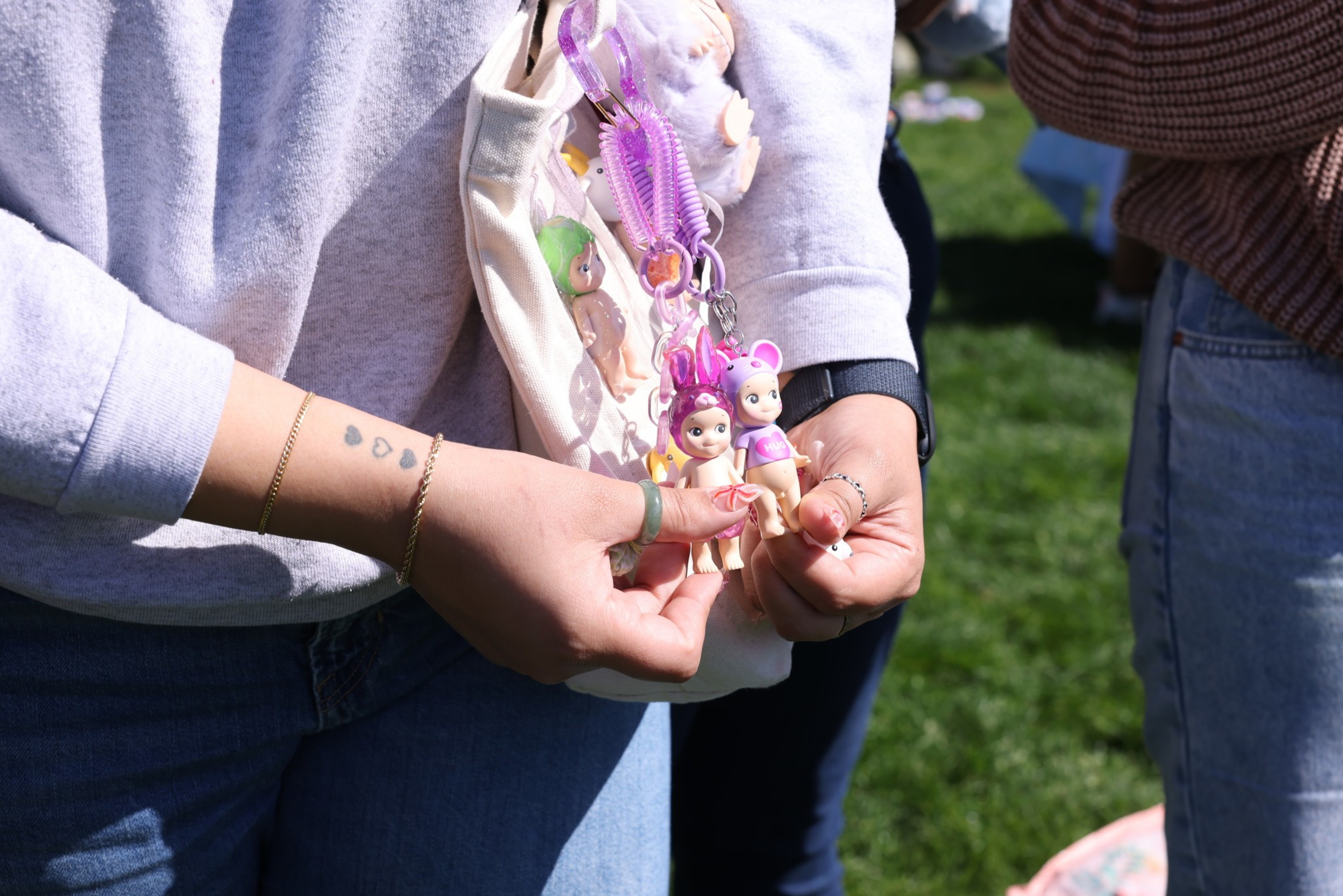 A person wearing jeans and a light sweater holds two small figurines. They have heart tattoos on their forearm and an open bag with more figurines visible.