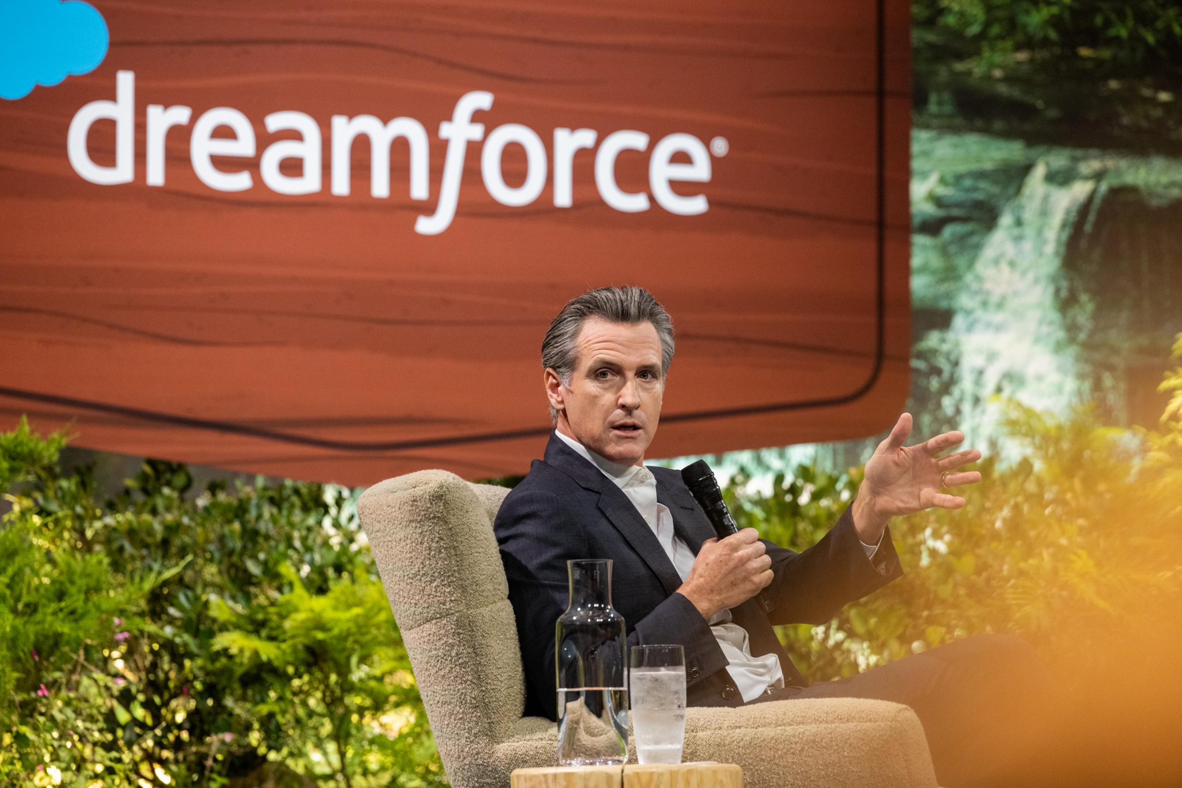 A man in a dark suit sits on a beige chair speaking into a microphone, with a glass of water and a carafe on a table nearby and a "dreamforce" sign above.