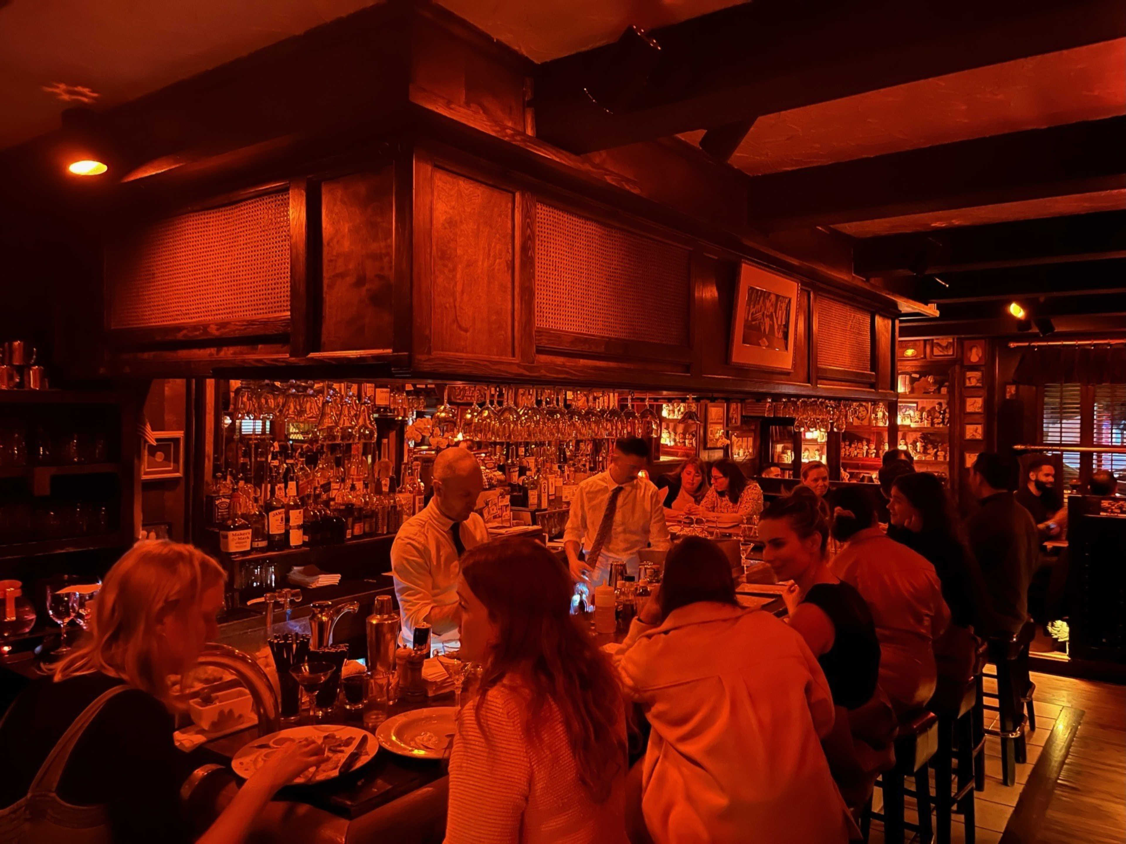A busy bar with warm orange lighting shows people seated at the counter and bartenders in white shirts serving drinks behind a wooden bar.