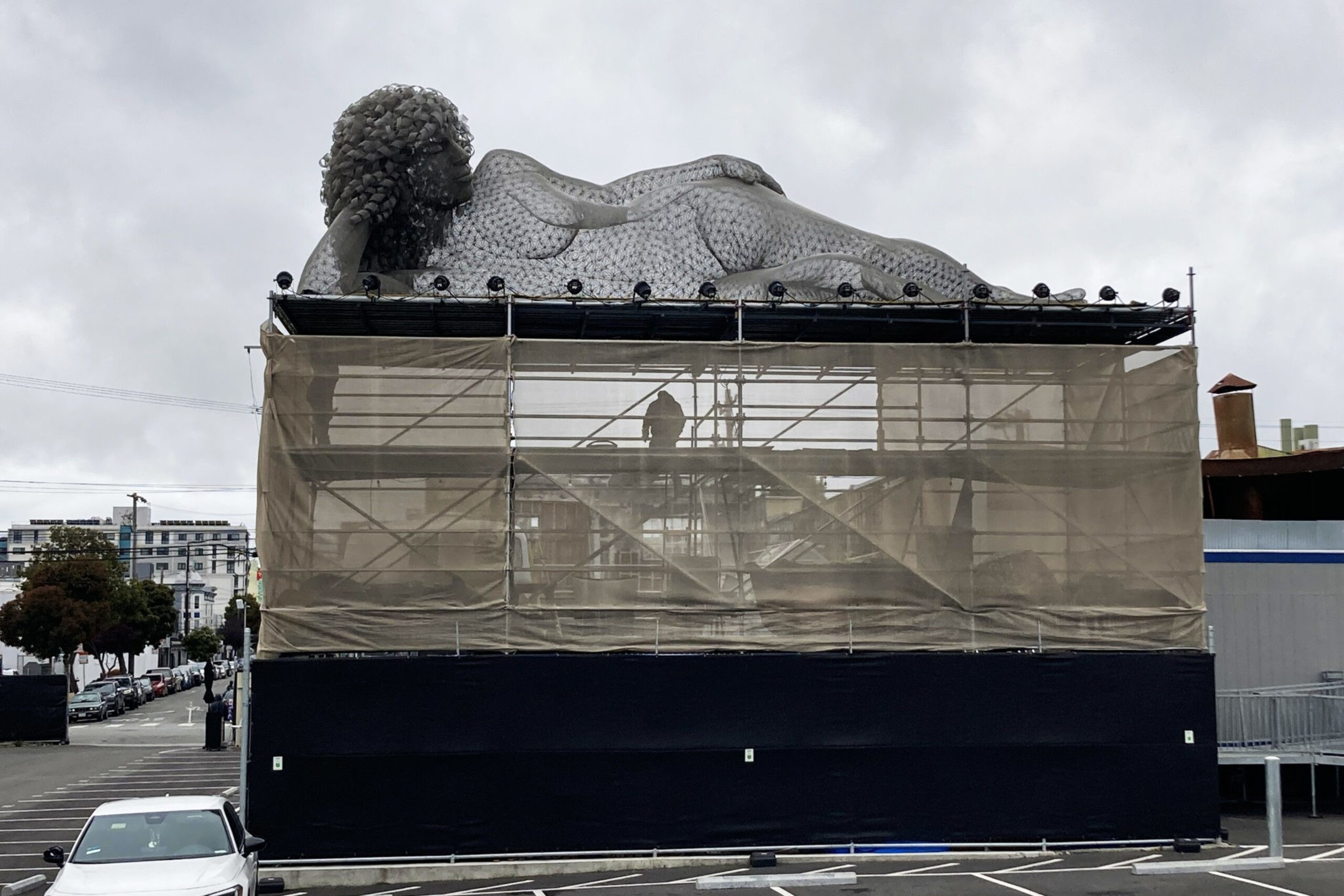 A large reclining woman sculpture with curly hair rests atop a scaffolding structure covered with mesh fabric in an urban parking area.