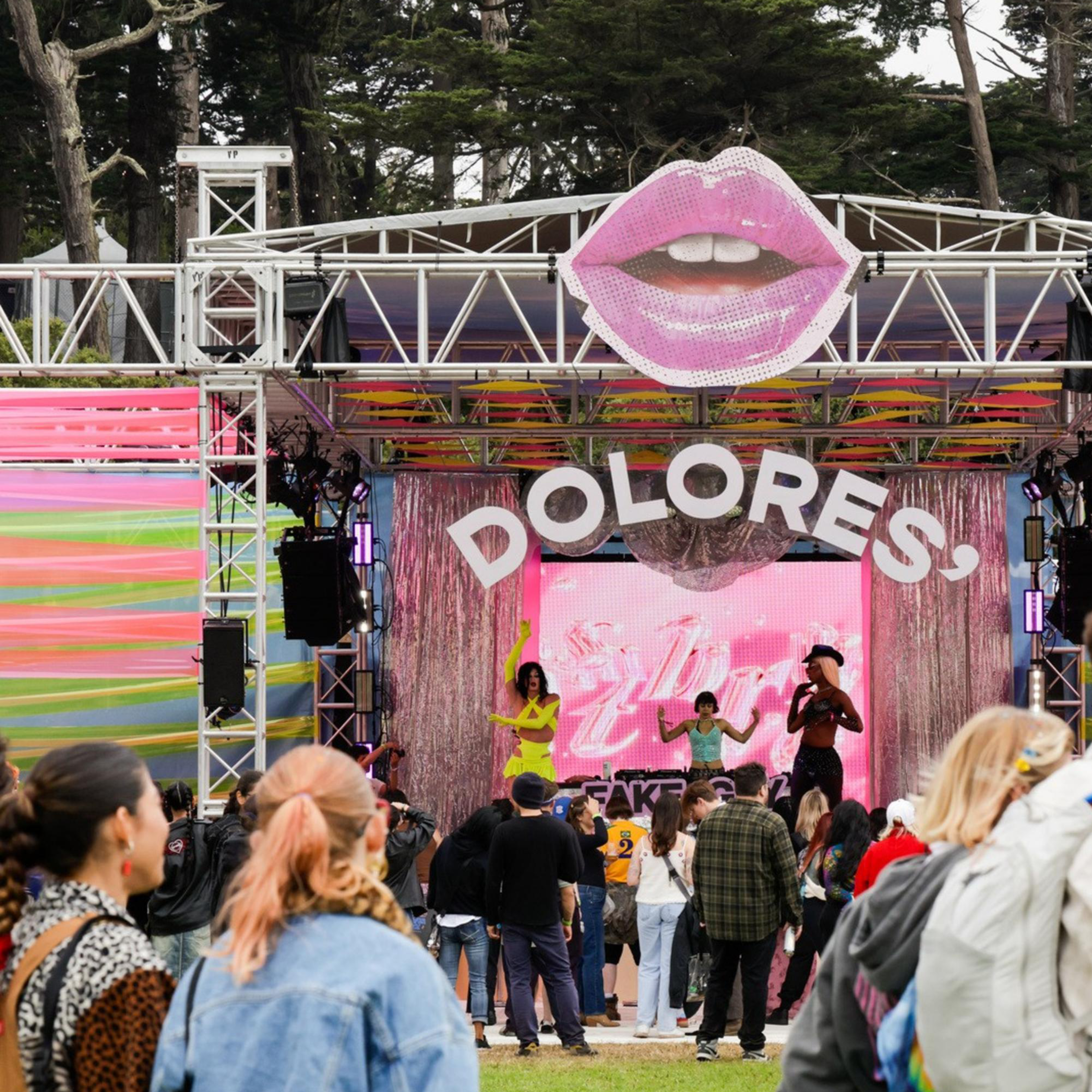 People gather at an outdoor festival stage with a large pink lips decoration above and the word &quot;DOLORES&quot; displayed, while performers entertain the crowd.
