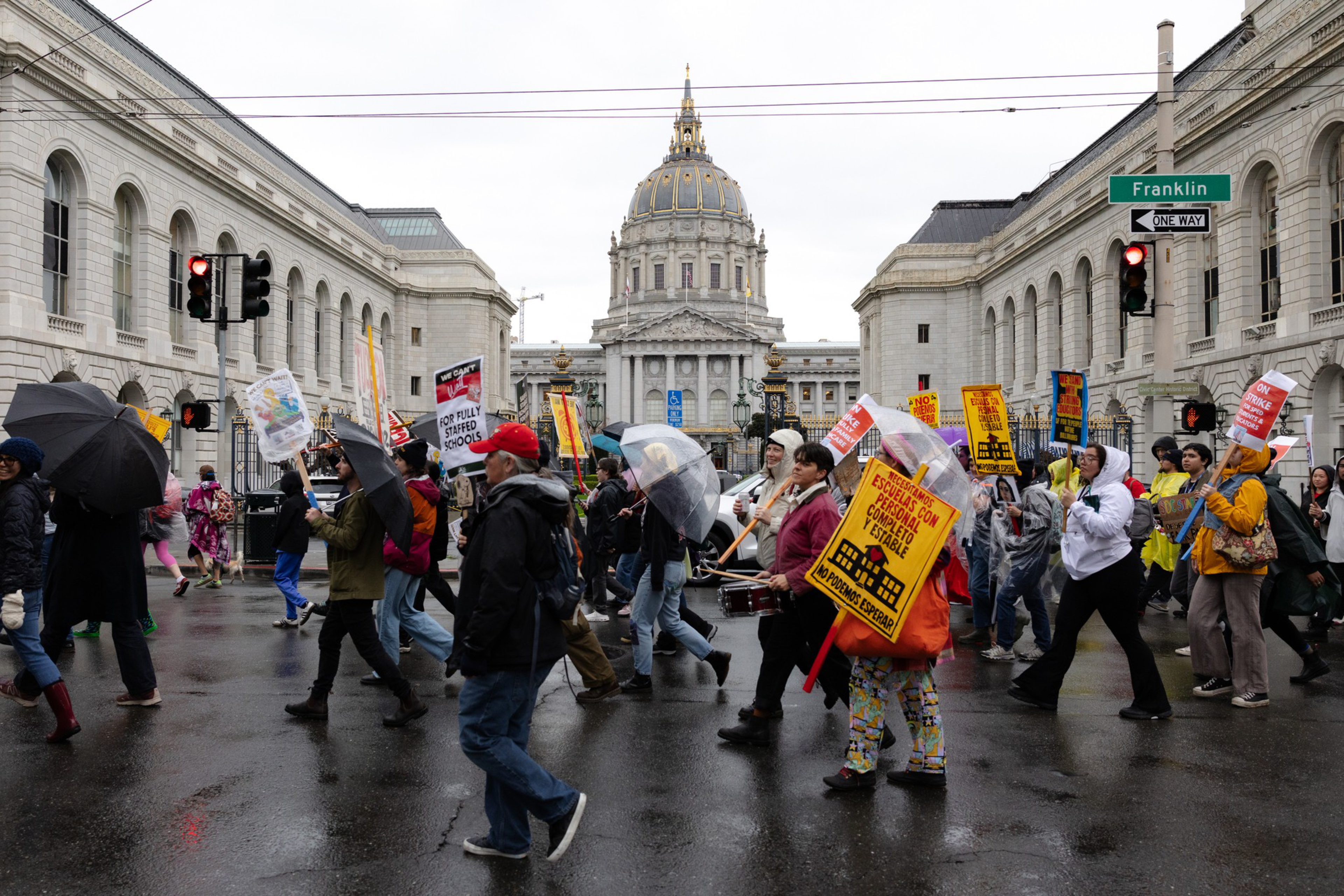 A group of people march on a wet street holding protest signs and umbrellas near a large domed building with "Franklin" street signs visible.