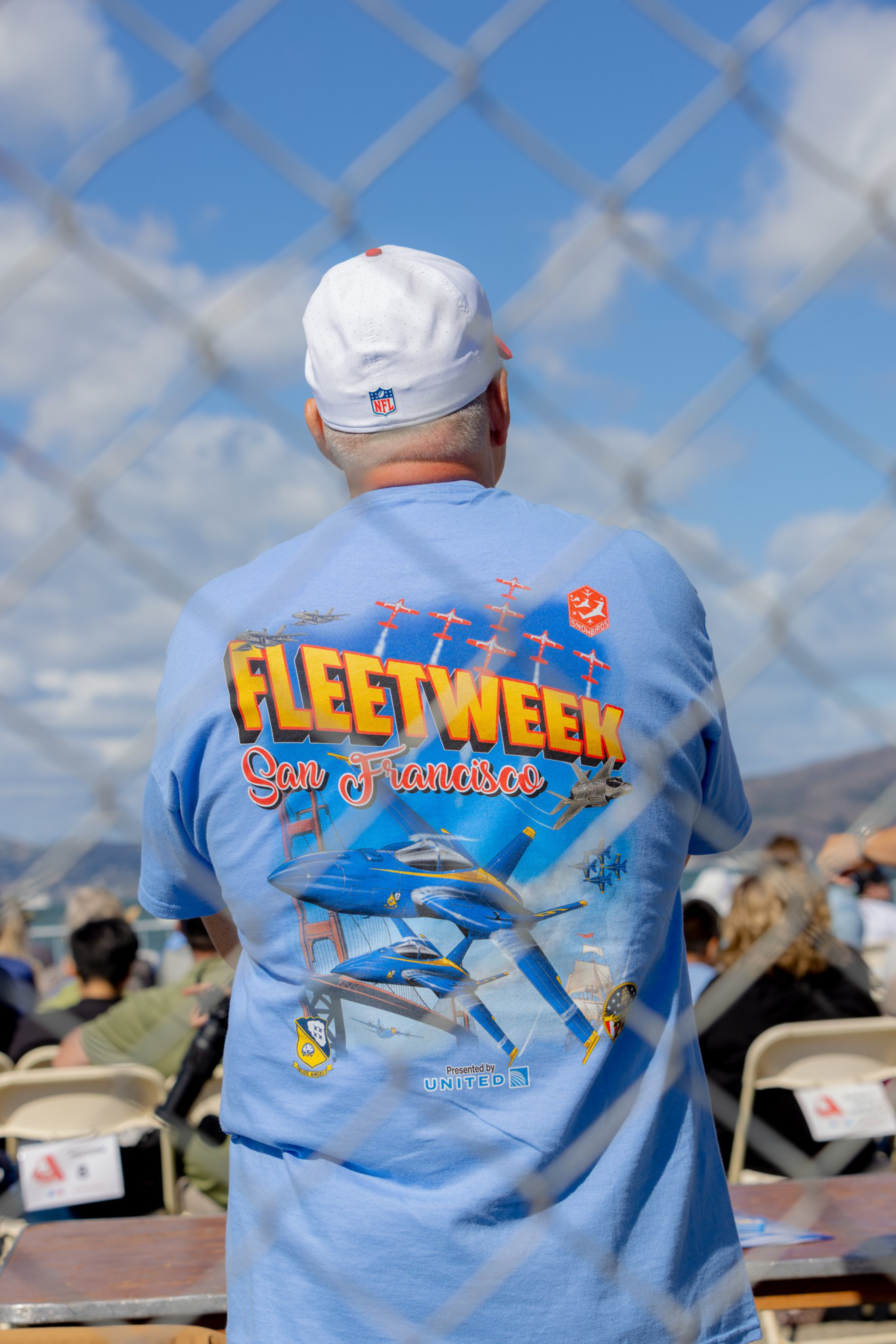 A person wearing a light blue “Fleet Week San Francisco” shirt featuring fighter jets and the Golden Gate Bridge sits behind a chain-link fence in an outdoor event.