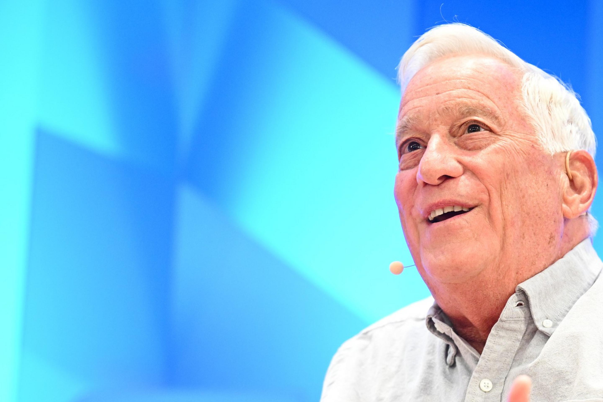 An older man with white hair is speaking animatedly on stage, wearing a light gray shirt. He's gesturing with his hands; a microphone is visible on his cheek.