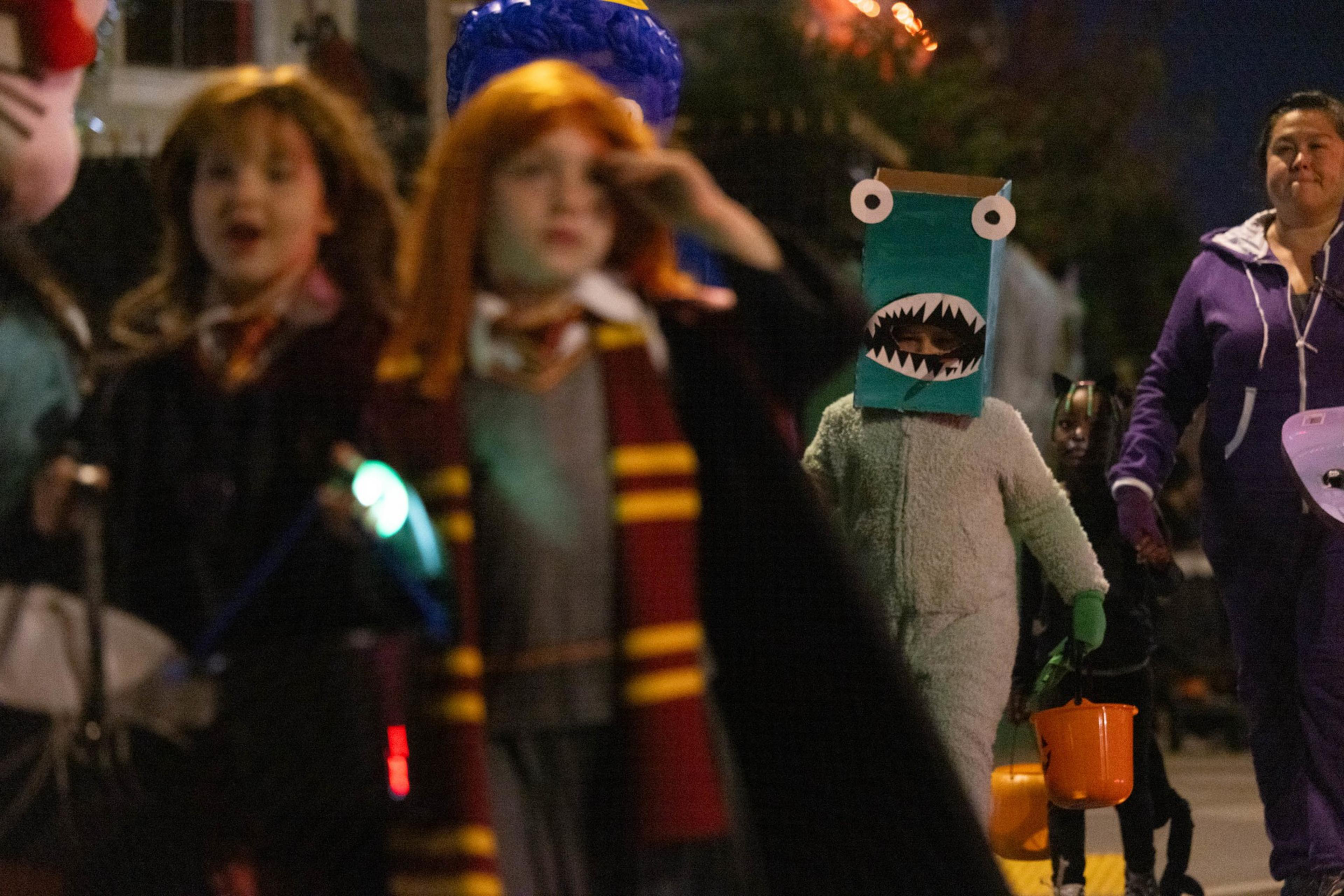 Children and an adult dressed in various costumes, including a shark mask and Hogwarts robes, walk outside at night during a Halloween event.