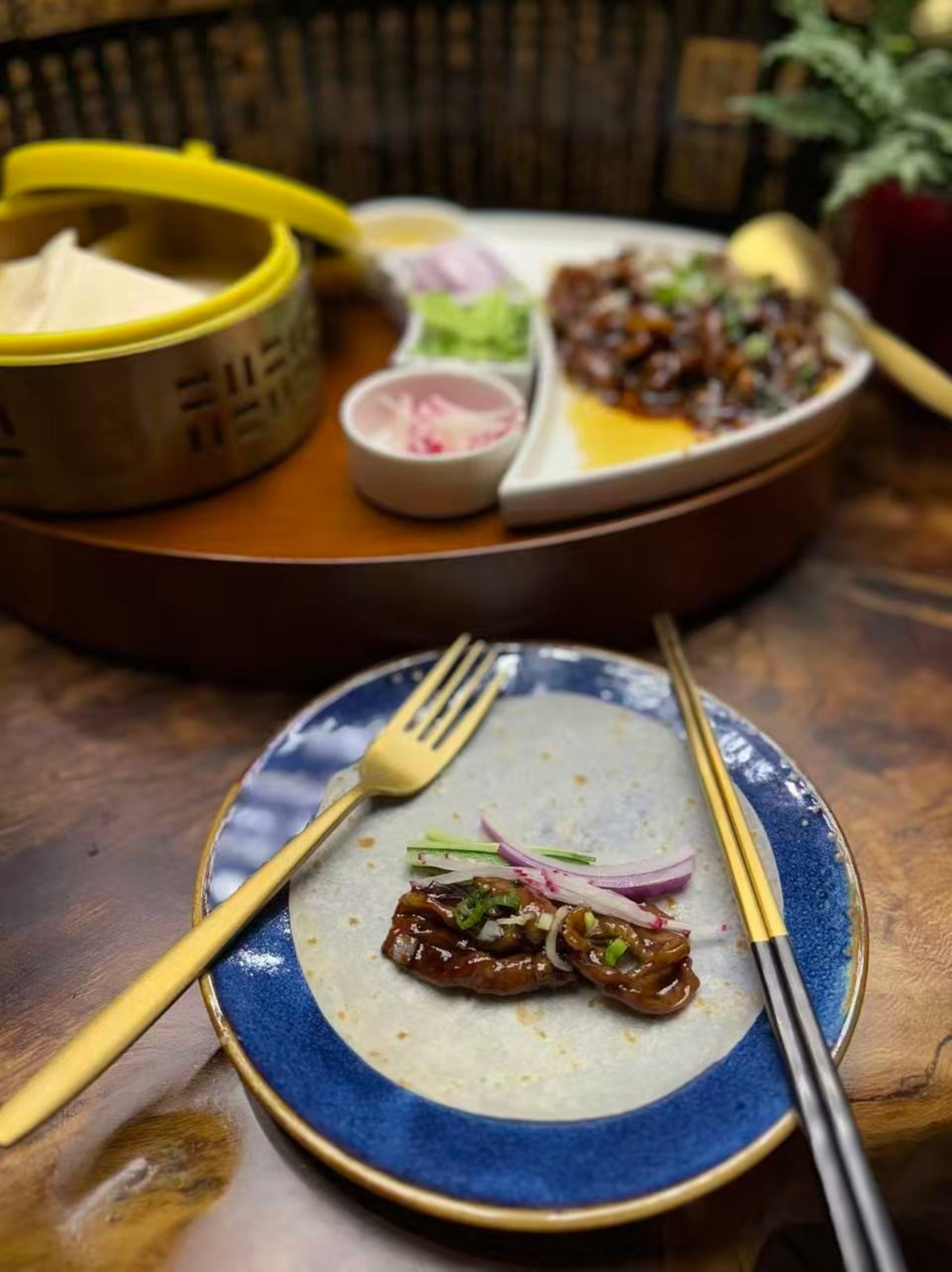 A tortilla on a blue plate holds grilled meat topped with sliced onions and herbs, with a gold fork and black-and-gold chopsticks placed beside it.