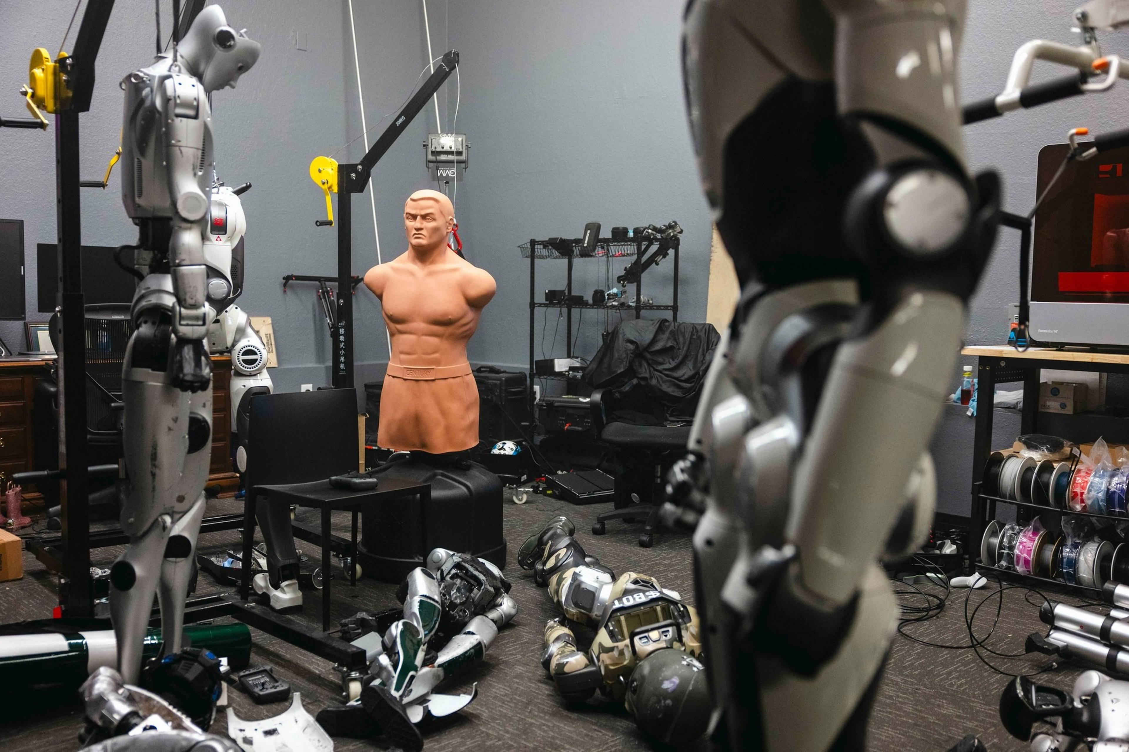 Humanoid robots stand and lie around a room with a human torso mannequin, shelves of equipment, and a computer workstation.