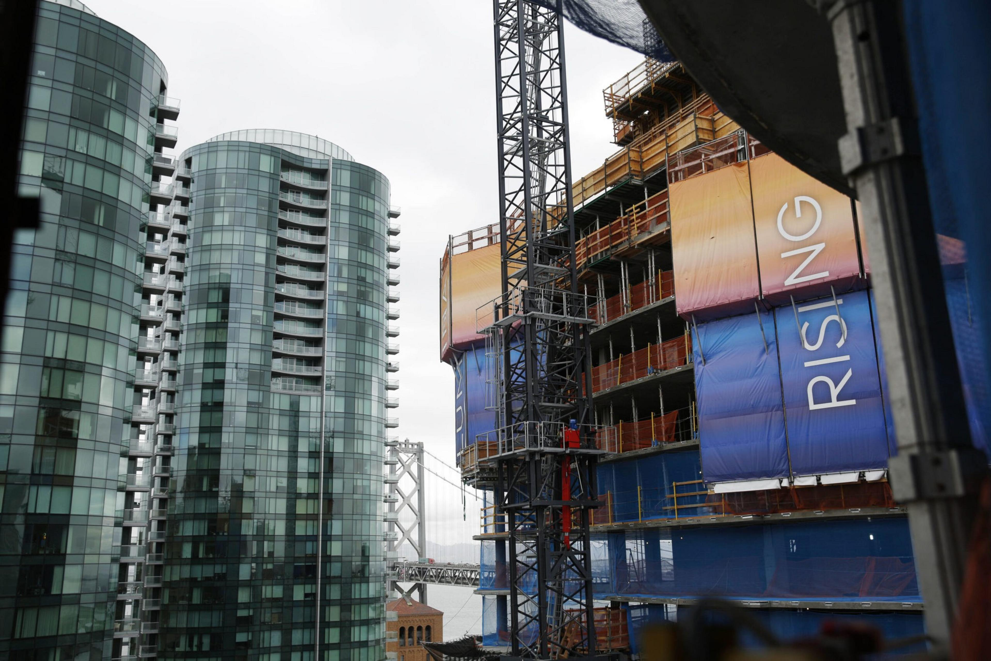 Modern glass apartment towers stand beside a construction site with a crane and colorful panels reading "RISING," with a bridge visible in the background.