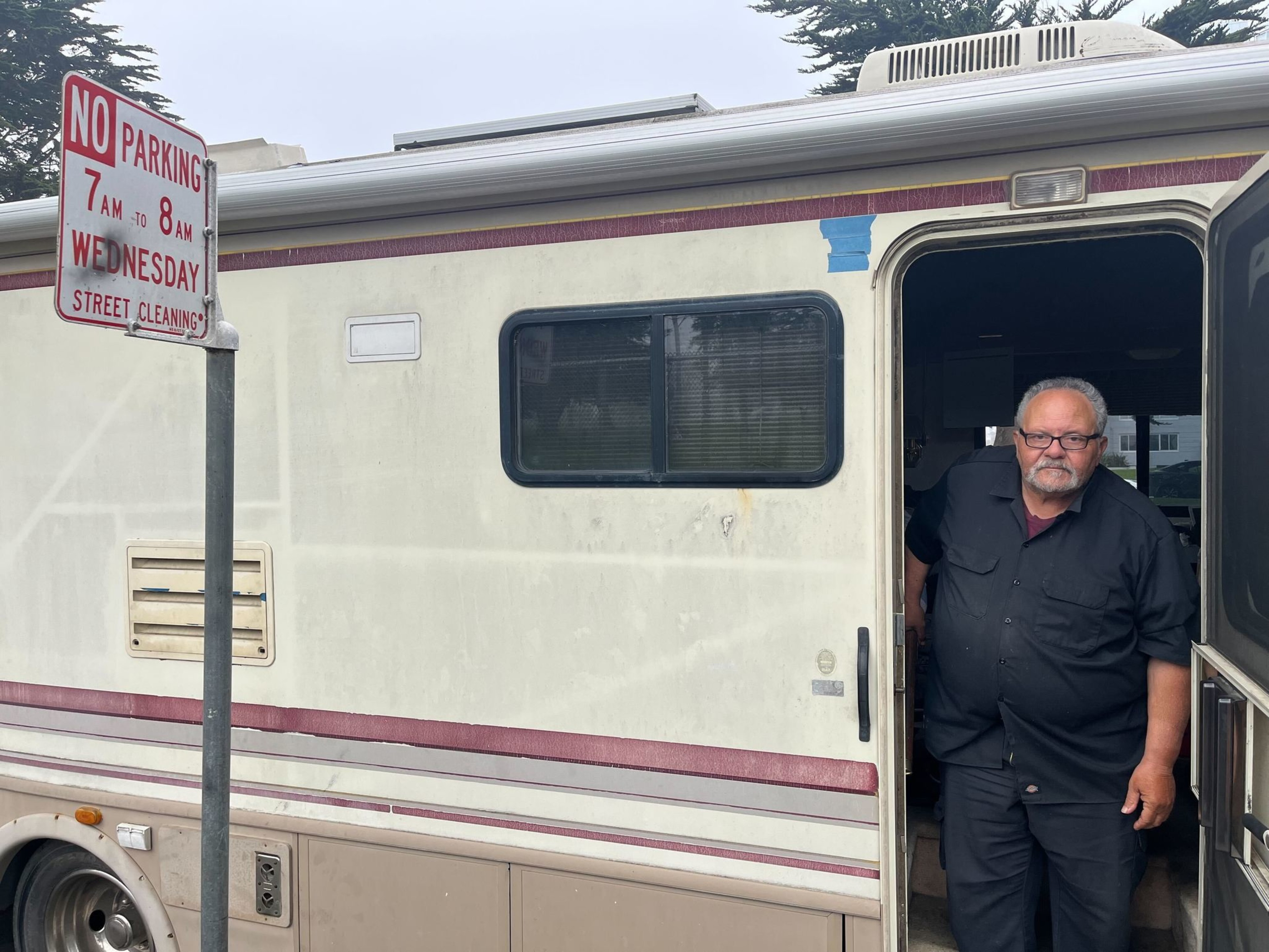 The image shows a man standing in the doorway of a parked RV. A street sign nearby indicates no parking during a specific hour for street cleaning.