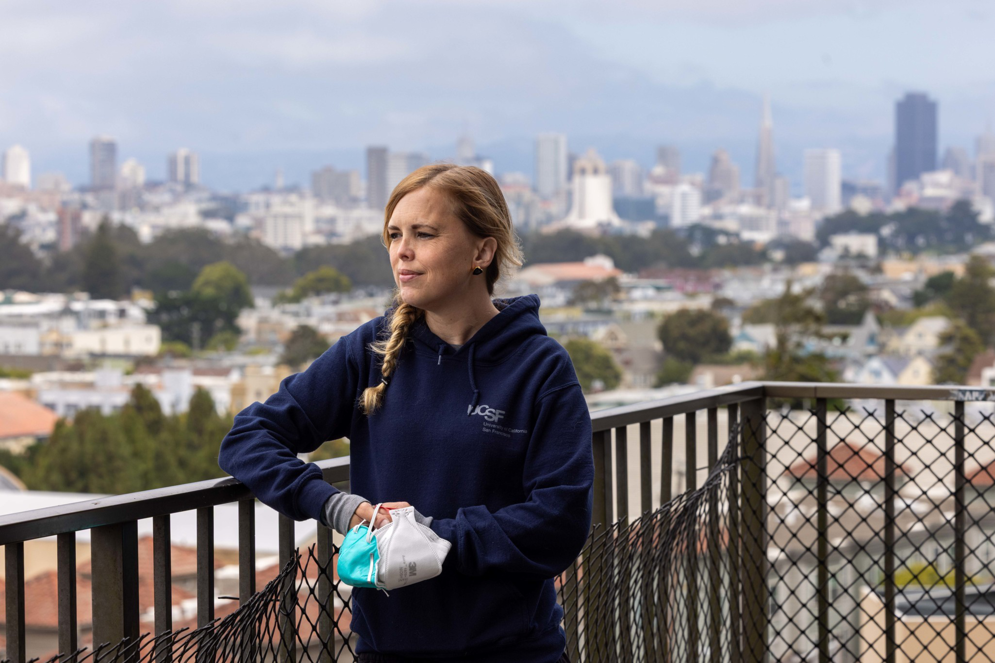 A woman in a navy UCSF hoodie stands on a balcony holding two face masks, looking thoughtfully toward a city skyline in the background.