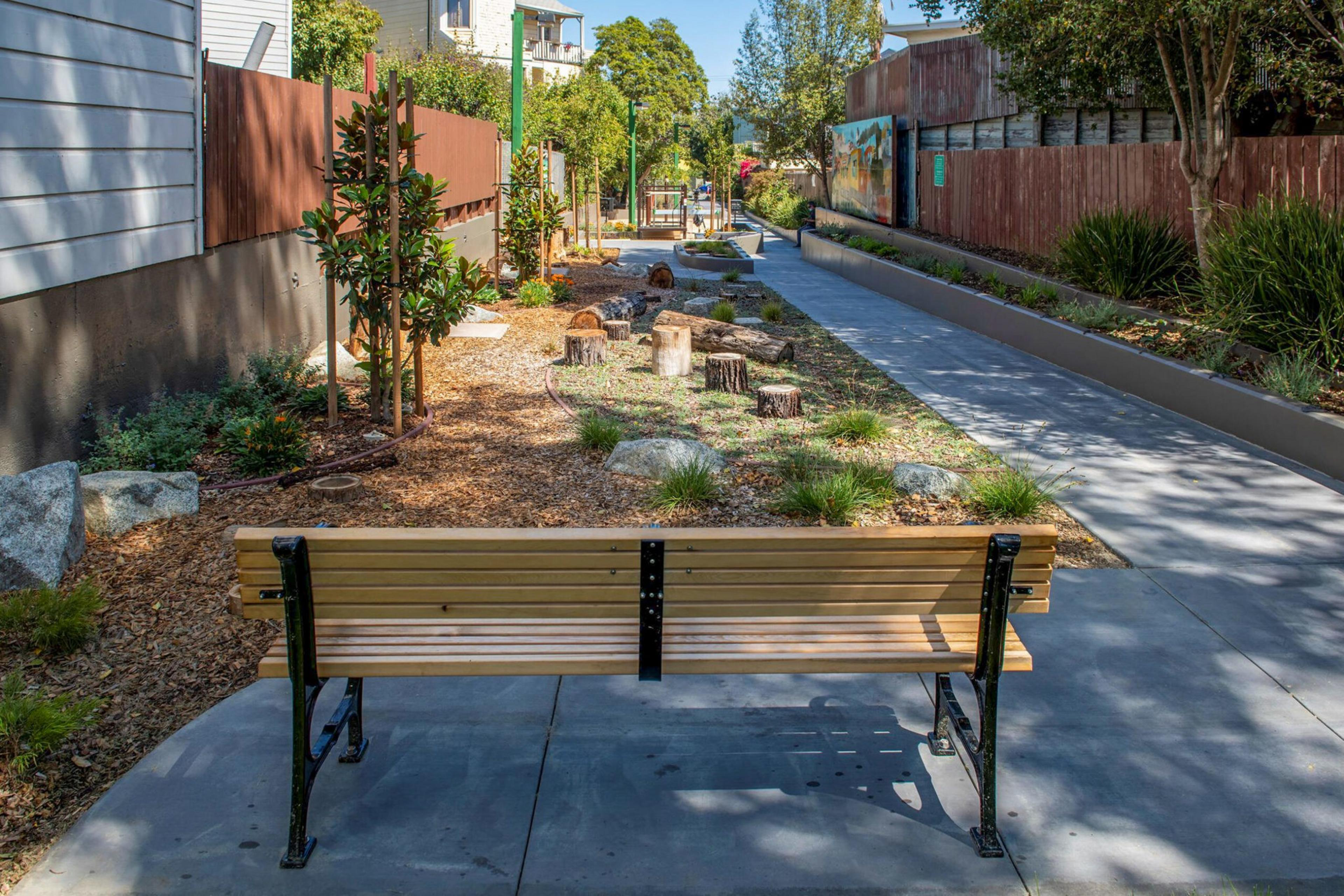 A wooden bench faces a landscaped area with tree stumps, rocks, and small plants, alongside a paved pathway and fenced yards.
