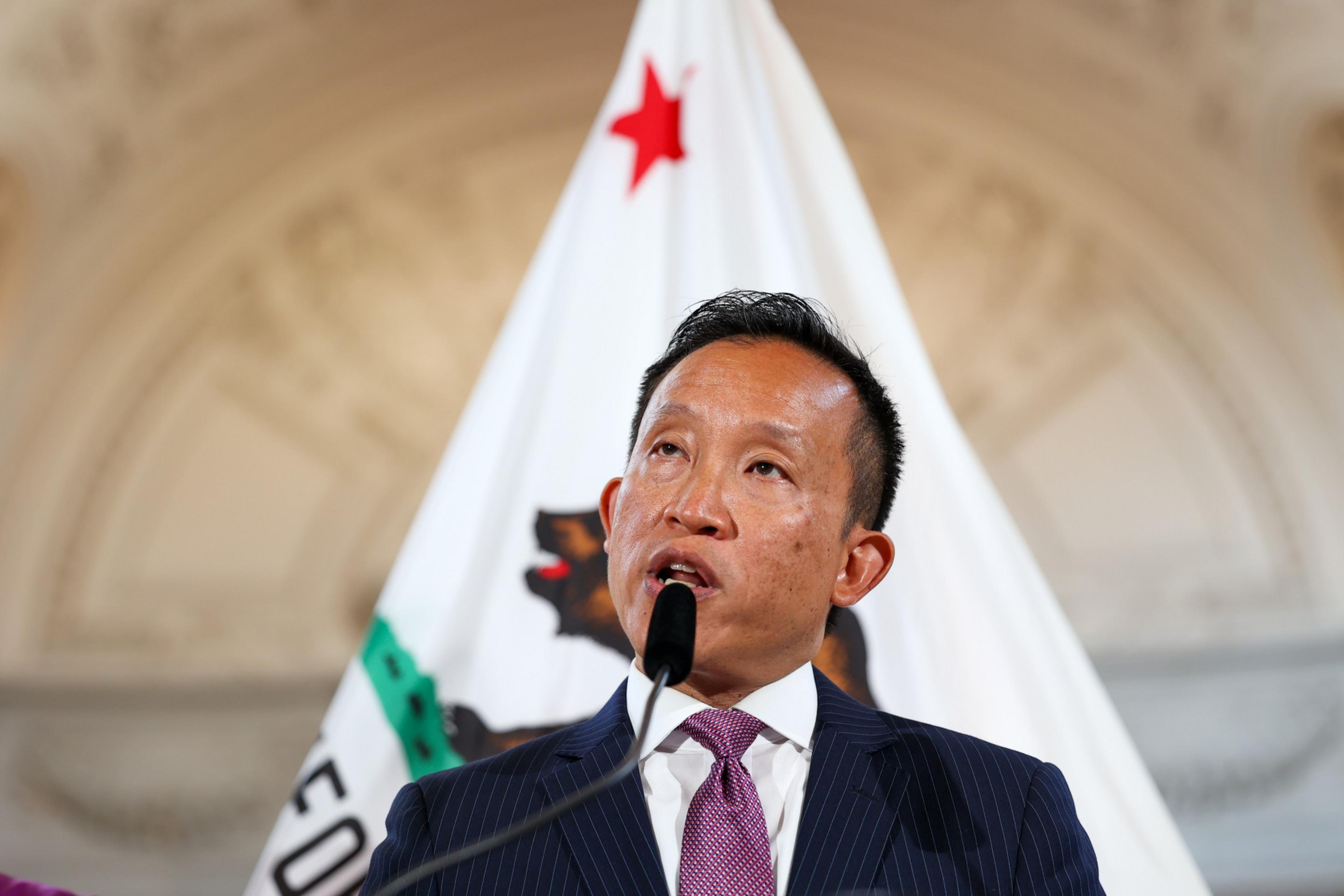 A man in a suit and purple tie speaks into a microphone, standing in front of a flag featuring a bear and a red star.