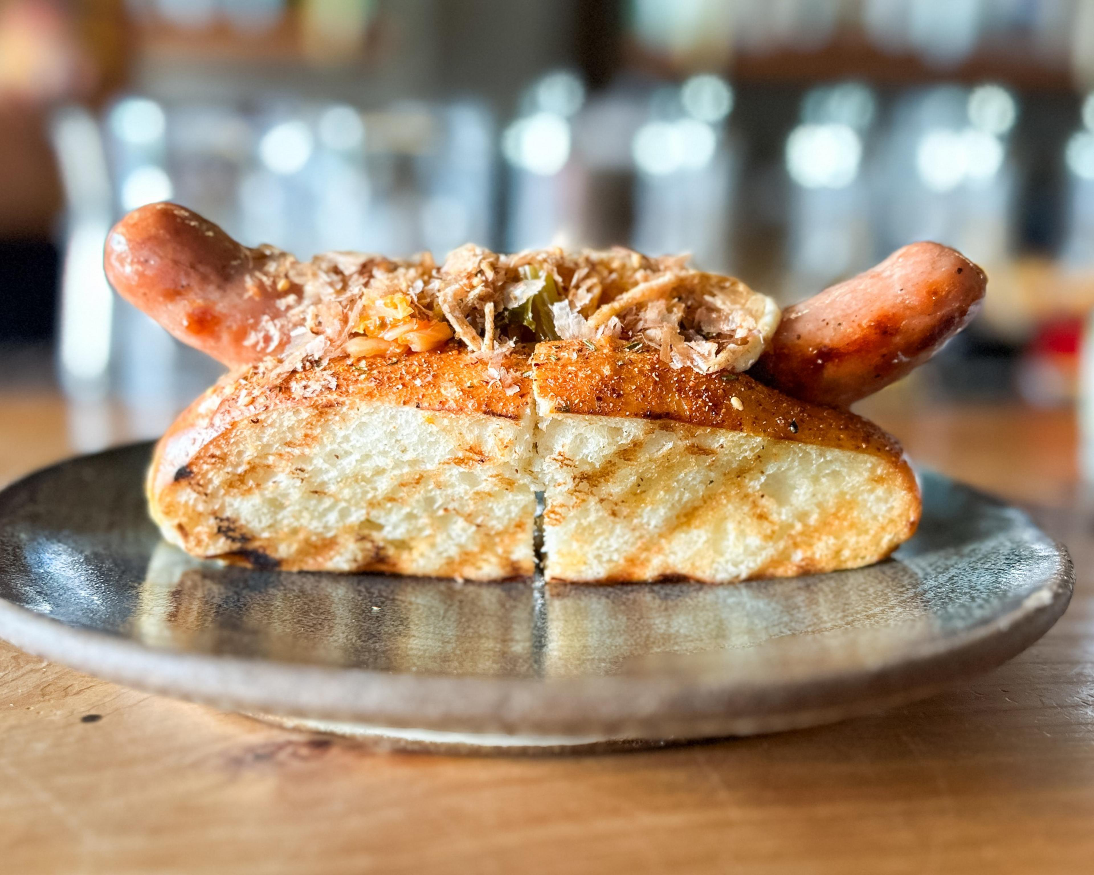 A hot dog on a toasted bun with two sausages, topped with shredded garnish and served on a black plate. It's placed on a wooden table.