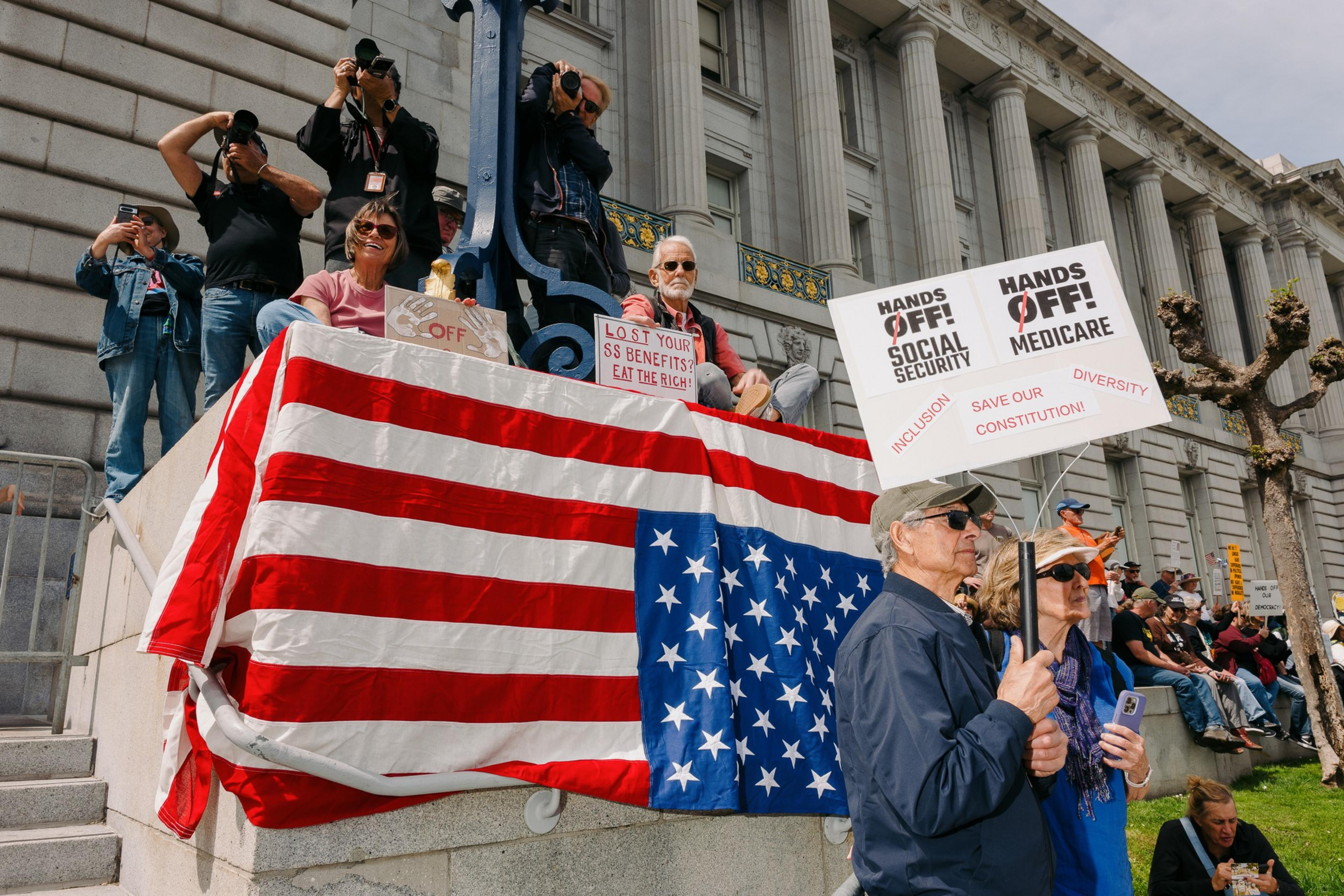 People are gathered outside a building, holding signs like &quot;Hands Off Social Security.&quot; An upside-down U.S. flag is displayed, symbolizing distress.