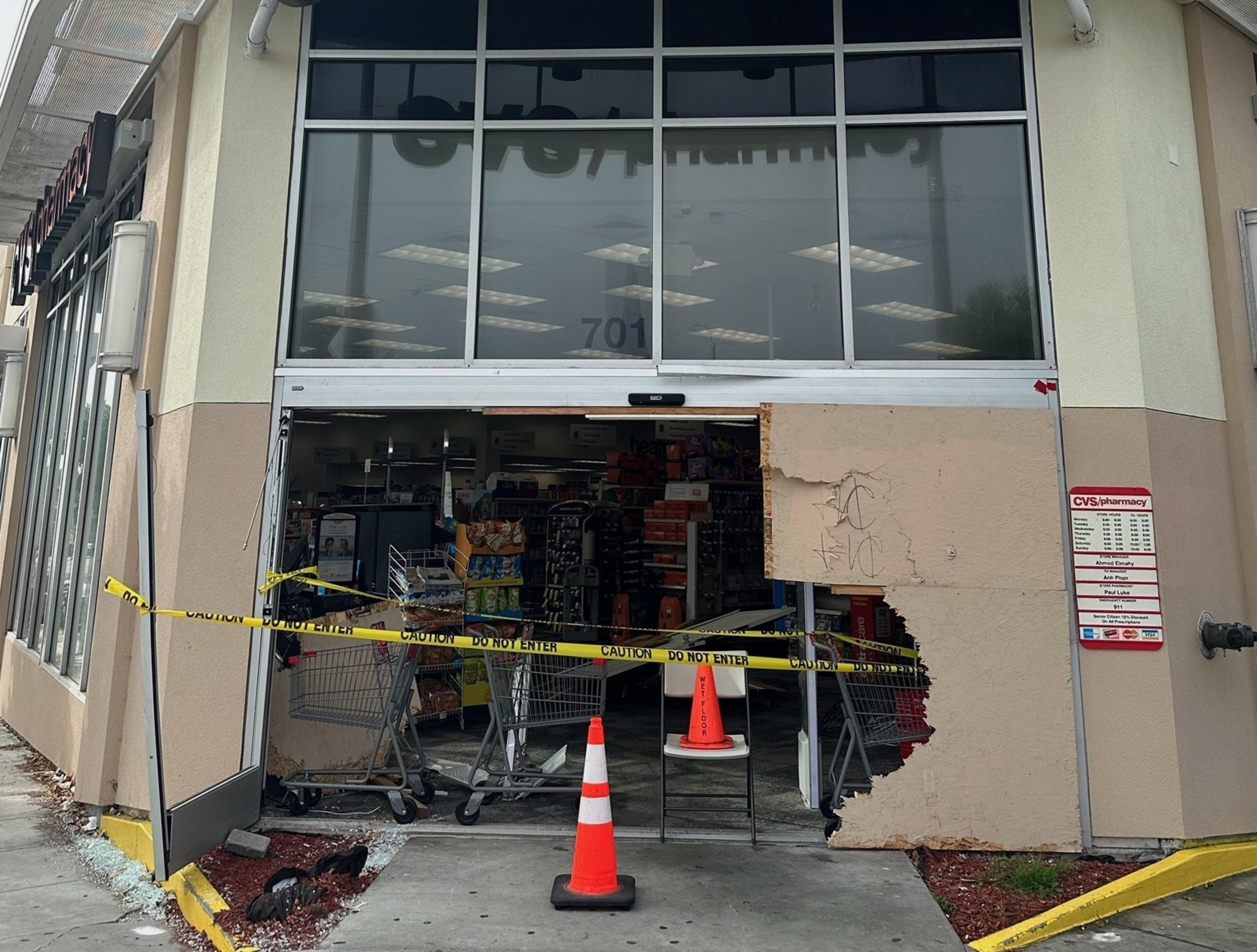 A damaged storefront with caution tape, a traffic cone, a damaged wall, and a view into the cluttered interior.