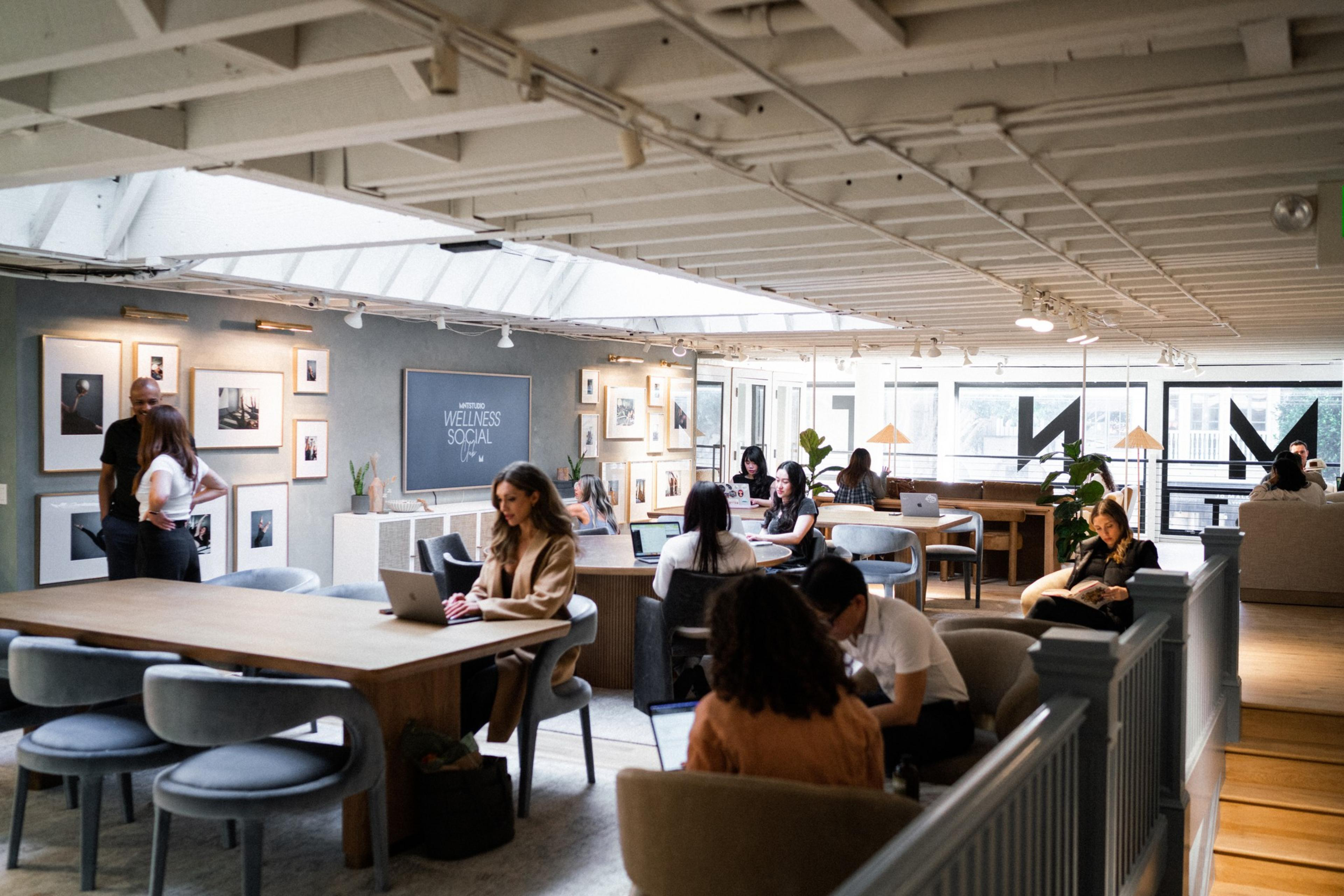 It's a cozy coworking space with natural light, people working on laptops at wooden tables, art on the walls, and plants scattered around the room.