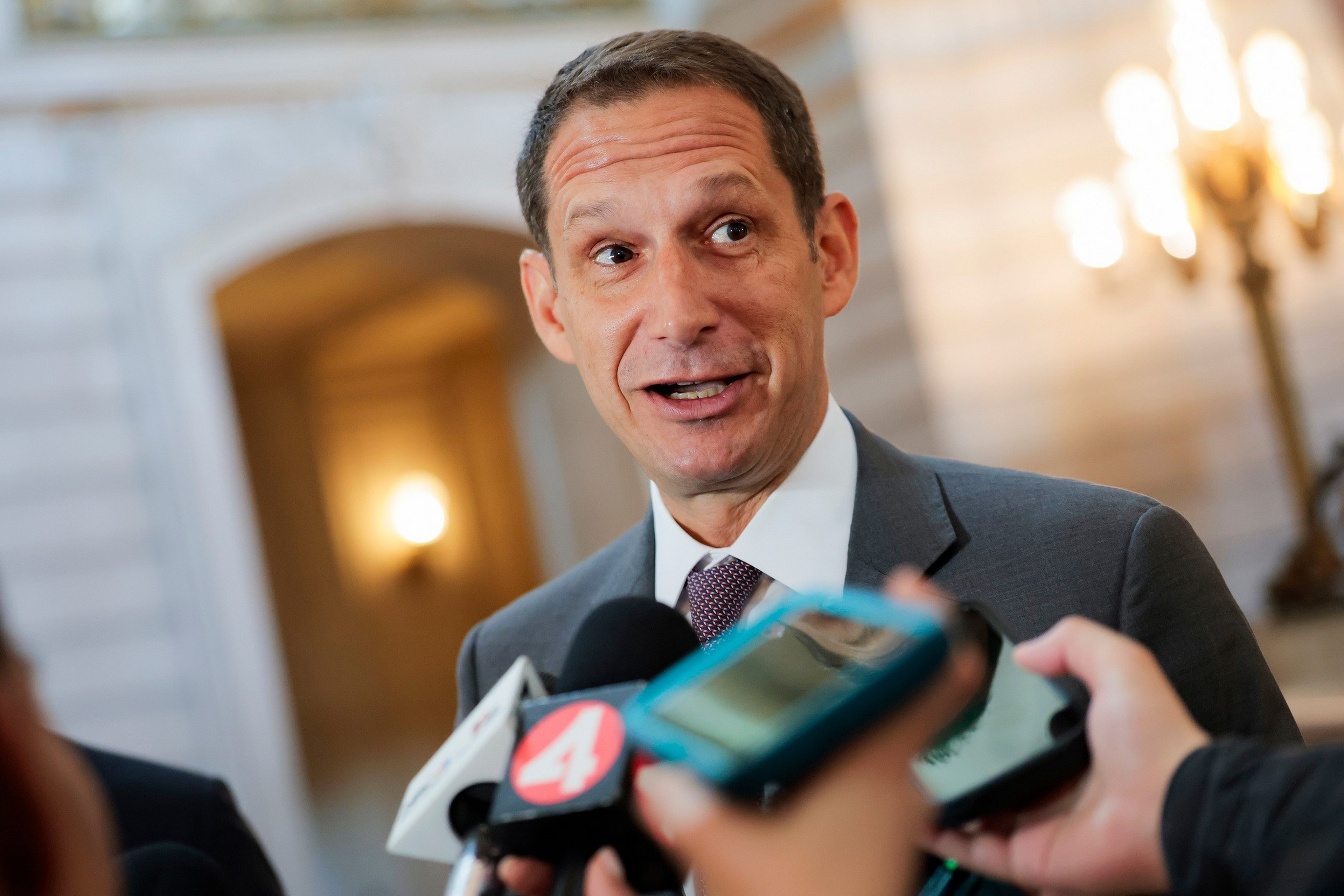 A man in a suit and tie speaks animatedly to reporters, with microphones and phones held up close to capture his statements.