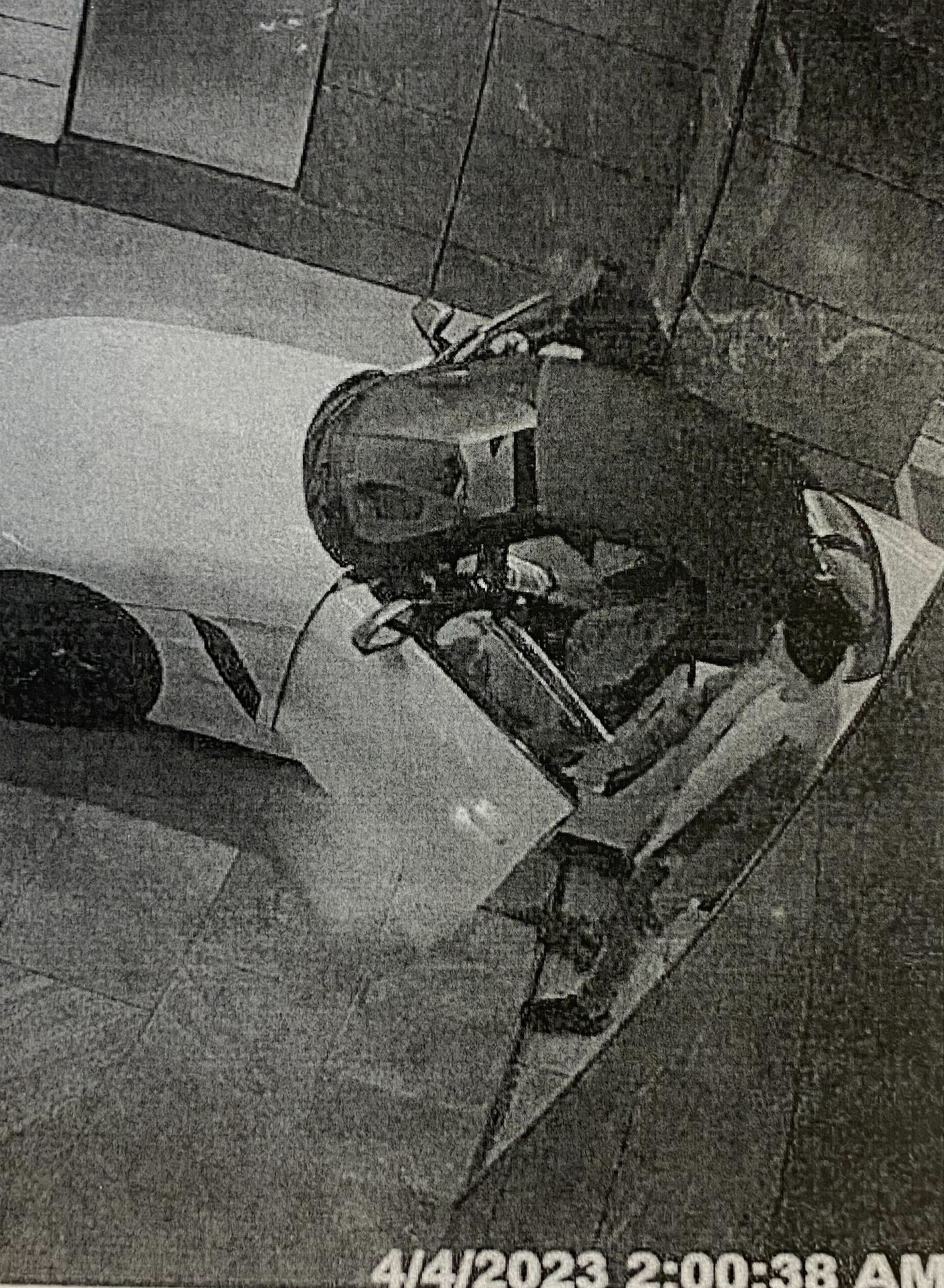 A security camera image shows a person entering a white car parked in a garage at 2:00 AM on April 4, 2023. The garage has tiled walls and flooring.