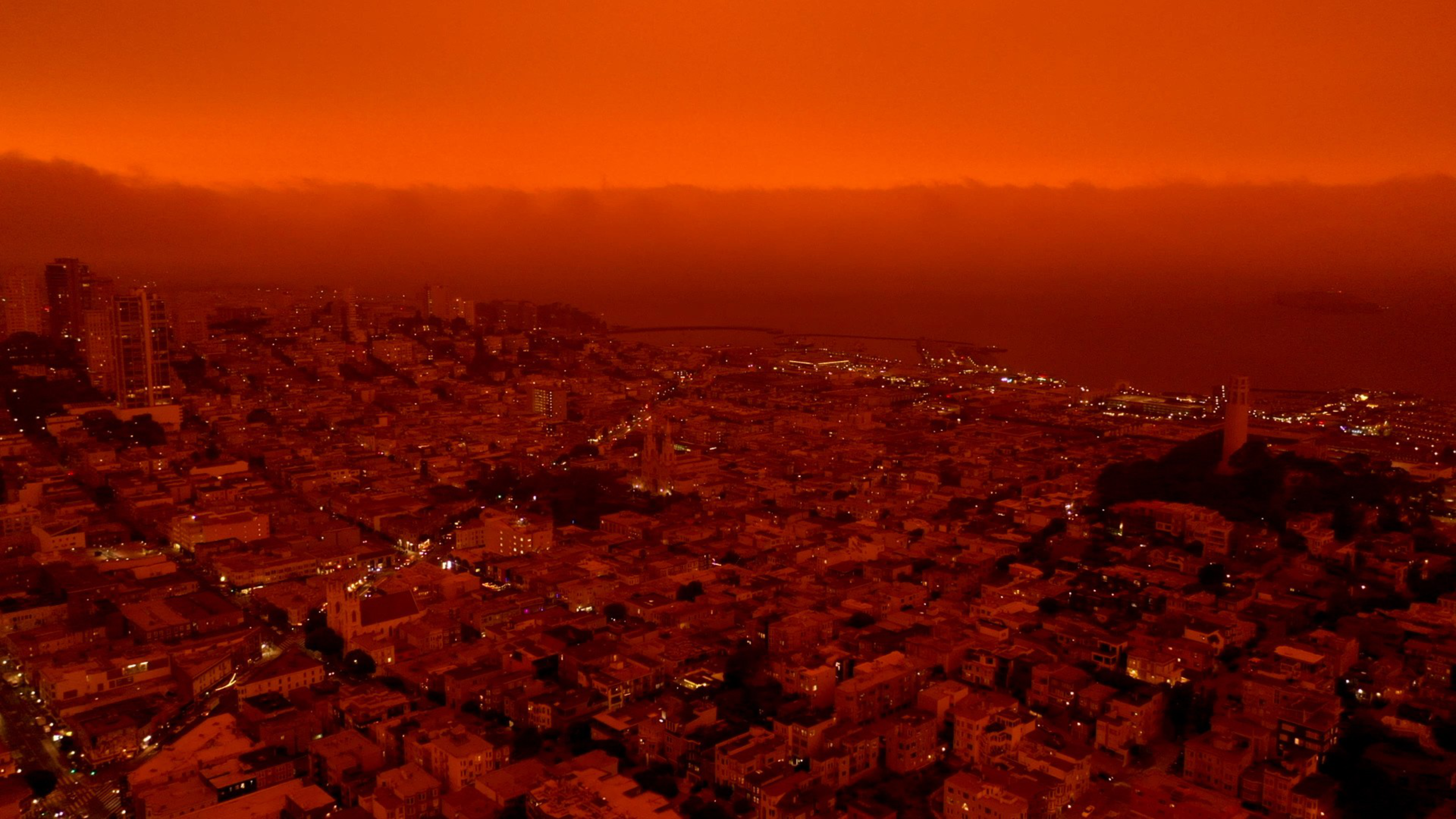 A cityscape under an intense red-orange sky, with illuminated buildings and streets stretching toward the horizon over water.