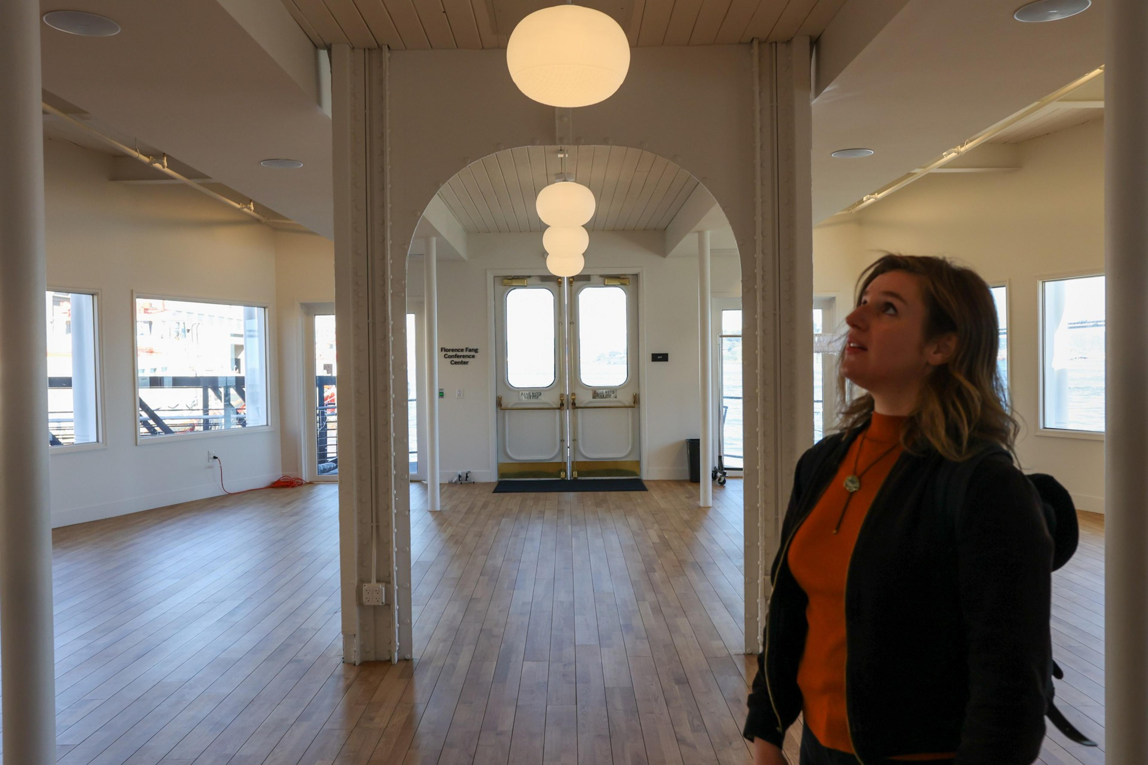 A woman in a black jacket and orange top looks up inside a spacious room with wooden floors, white walls, large windows, and round ceiling lights.