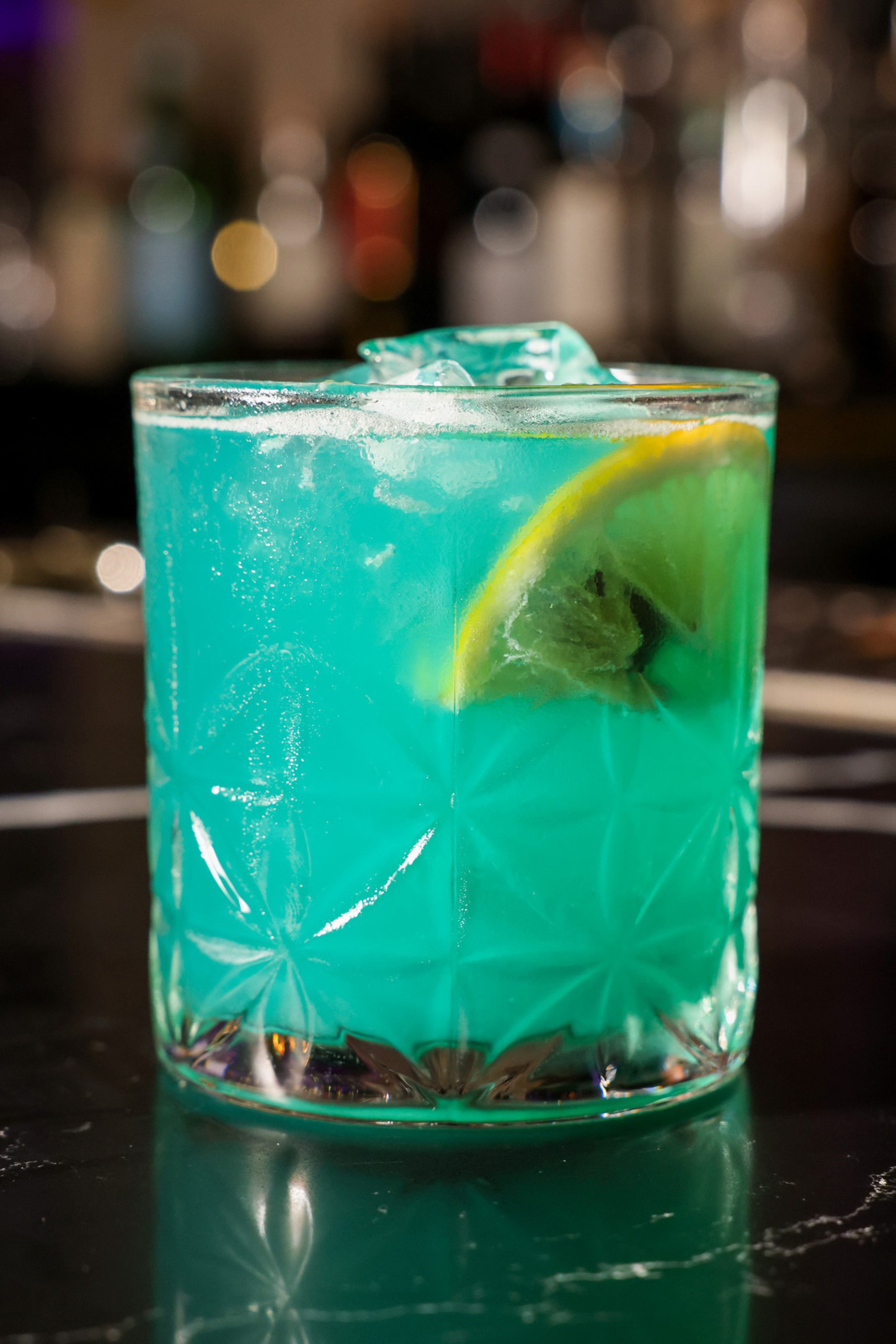 A bright blue cocktail in a patterned glass with ice and a lemon slice, set on a dark, glossy surface with blurred lights in the background.