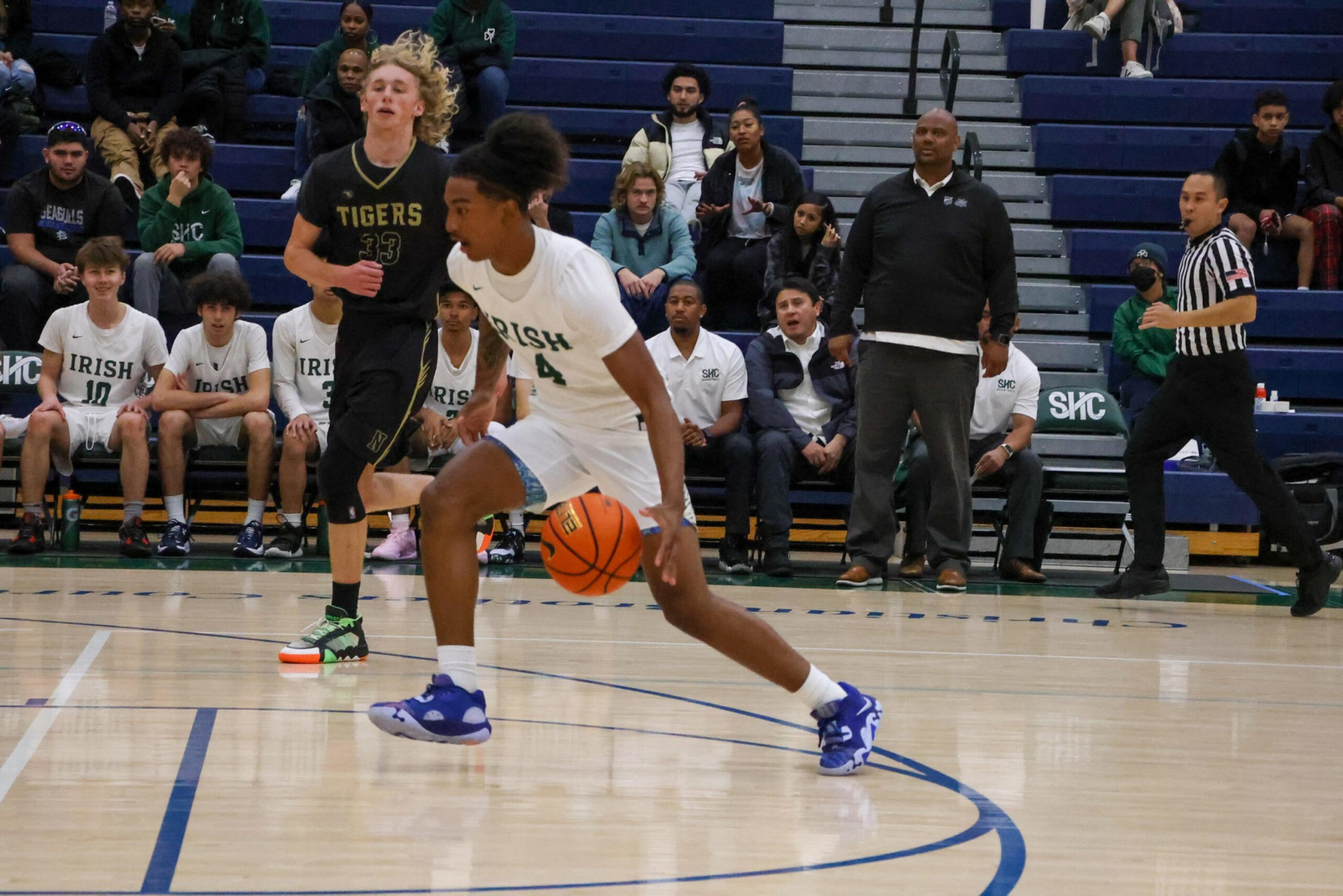 A basketball player wearing a white uniform dribbles the ball on the court while an opponent in a black "Tigers" uniform runs behind him.