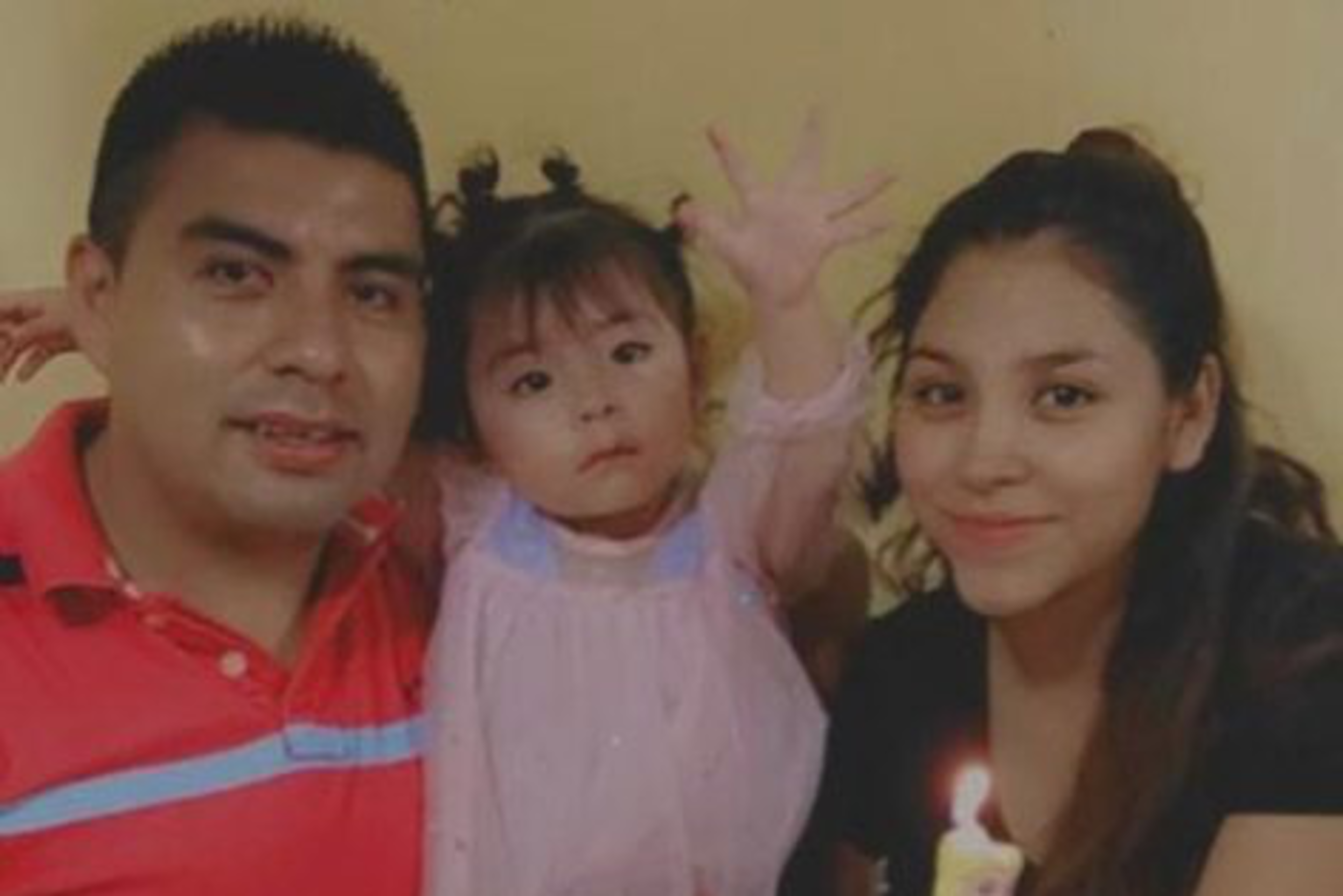 A family photo with a man, toddler, and woman; the child, raising her hand, holds a unicorn toy.
