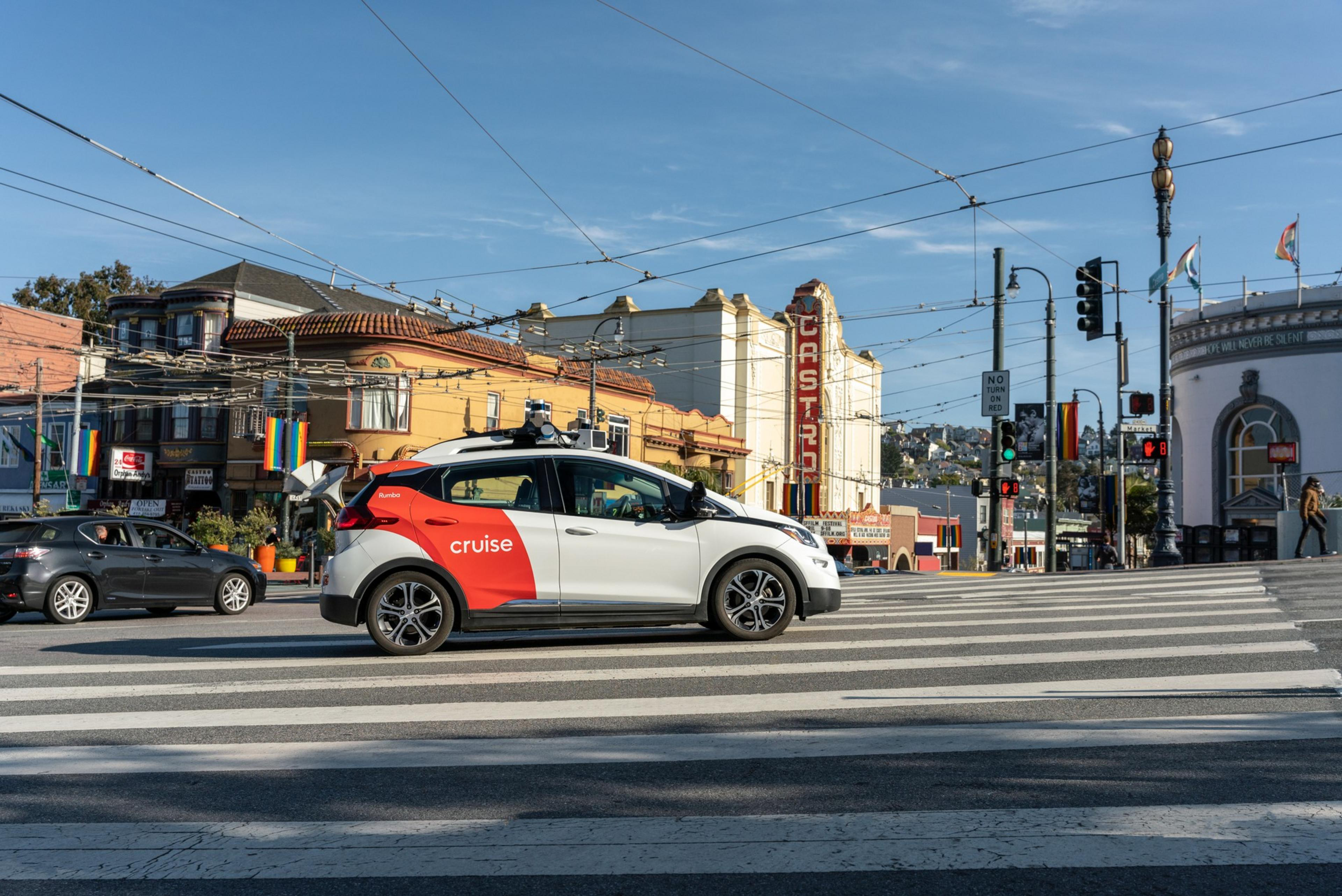 A white and red Cruise autonomous car waits at a crosswalk in a sunny urban area with vintage buildings and various traffic lights.
