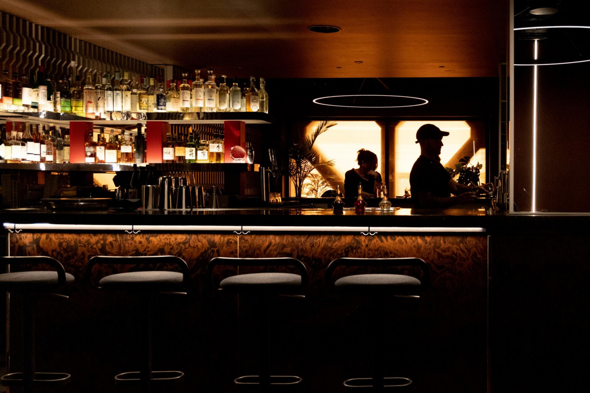 A dimly lit bar with four empty stools lined up, bottles on shelves behind the counter, and two silhouetted figures working inside.