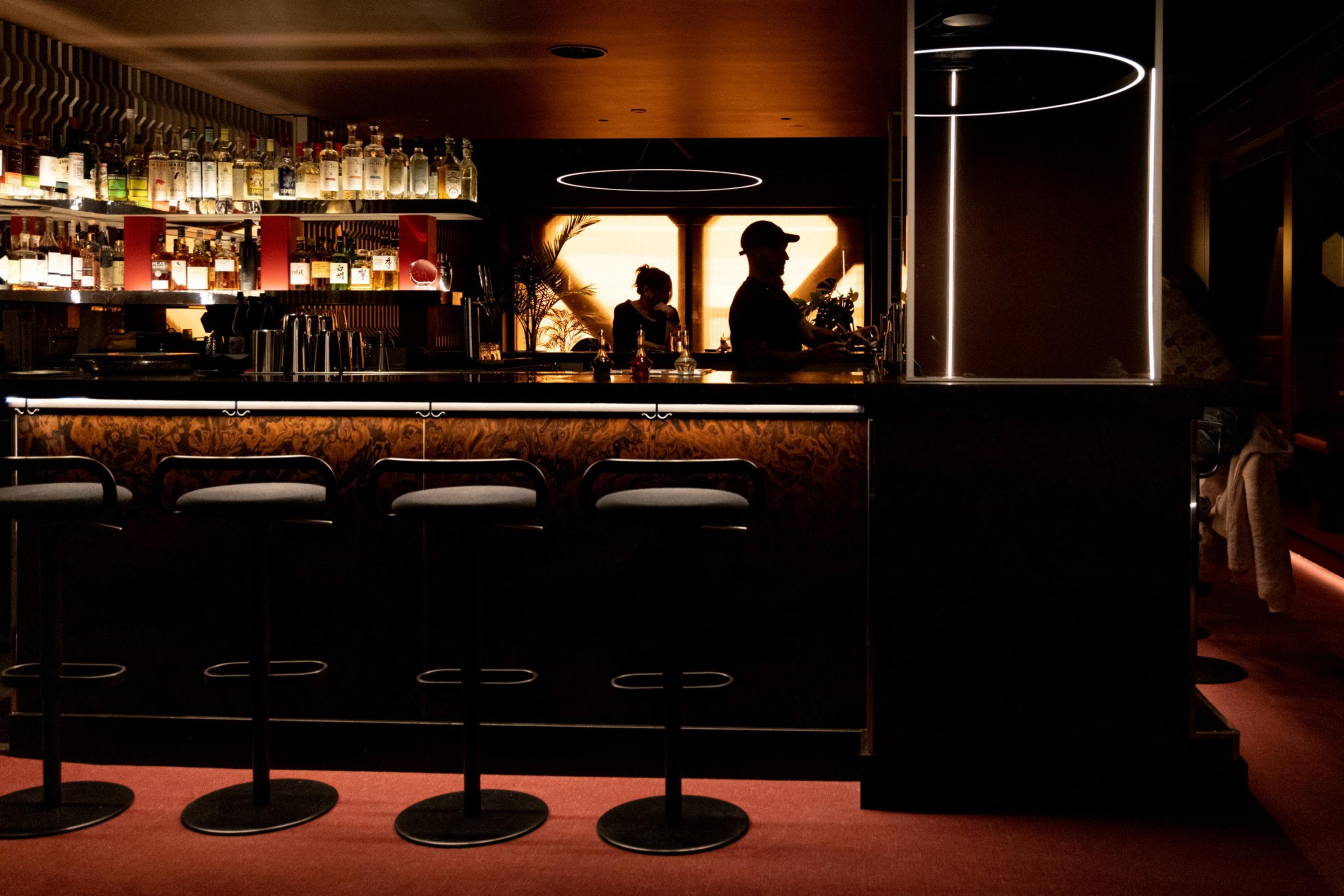 A dimly lit bar with four empty stools lined up, bottles on shelves behind the counter, and two silhouetted figures working inside.